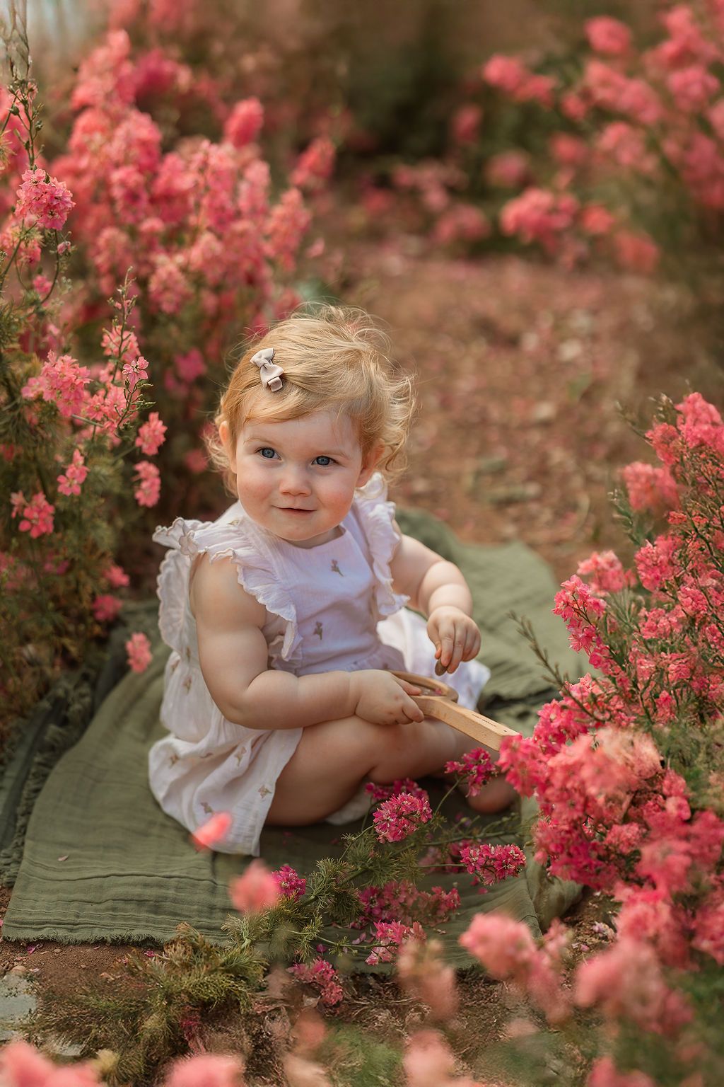 Adorable baby girl in white dress sitting among pink flowers outdoors.