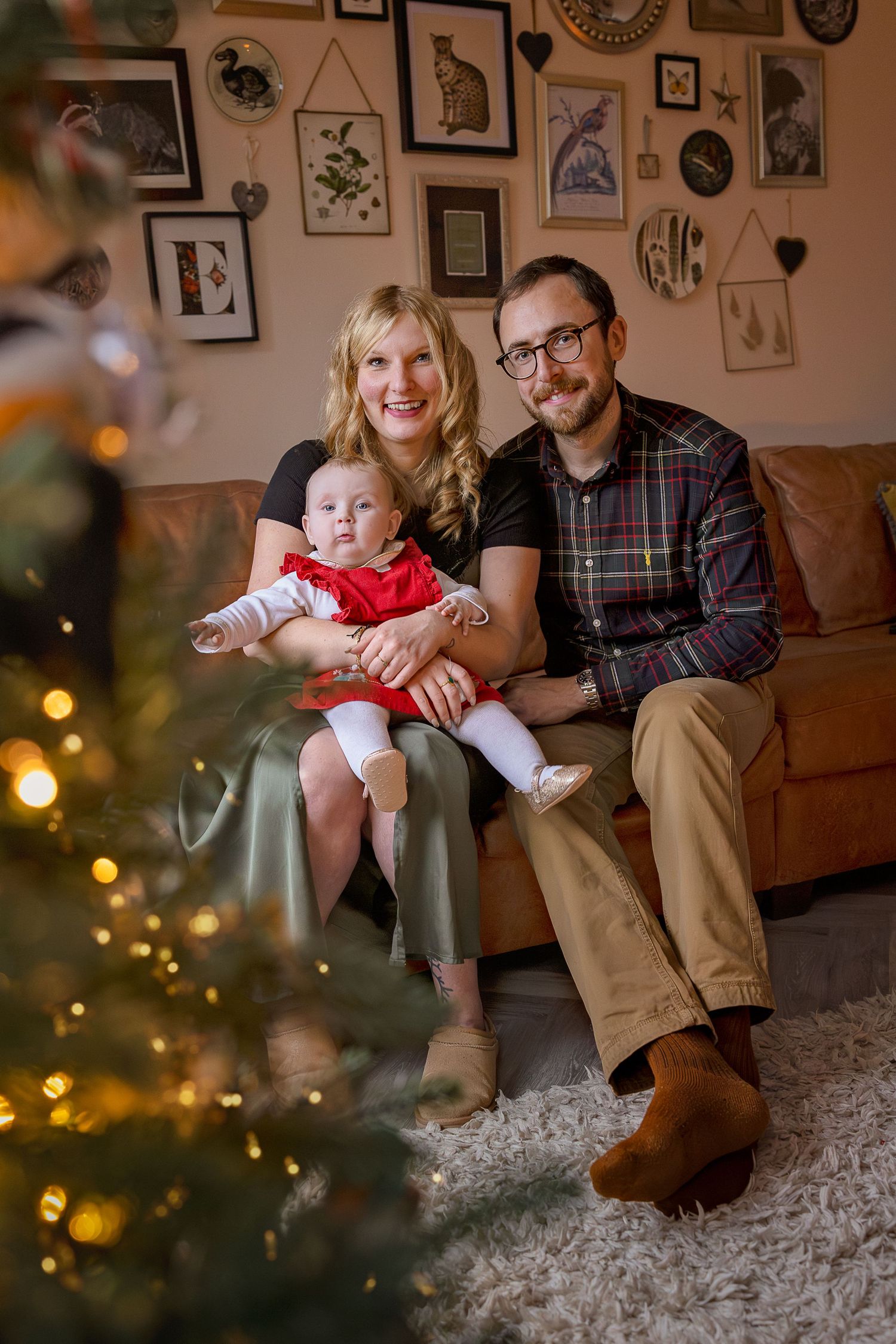 Family of three posing by Christmas tree on sofa, mom holding baby in red dress, dad in plaid shirt, festive home decor.
