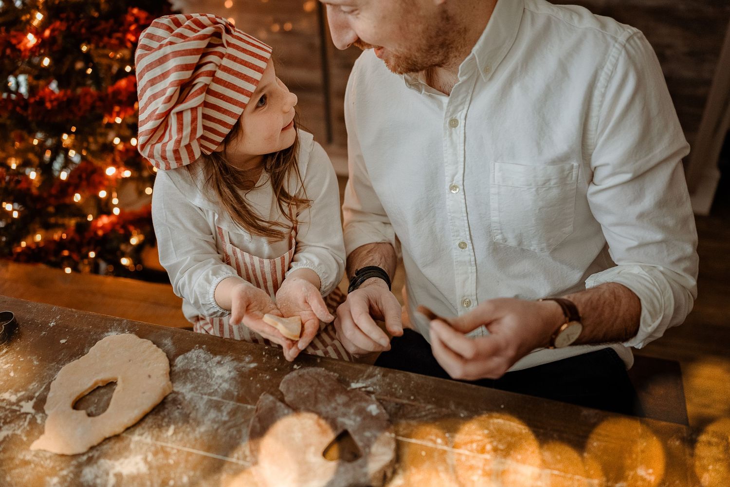Father and young daughter in a striped elf hat cut out Christmas cookie dough together near a glowing, decorated holiday tree.