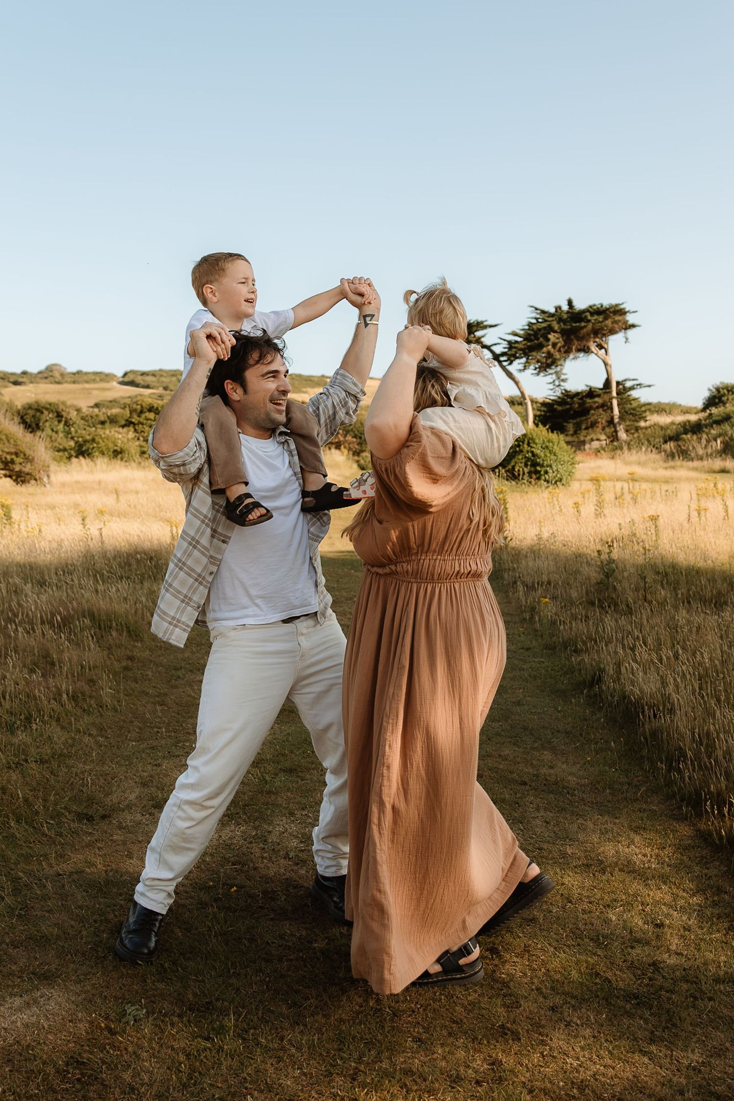 A couple enjoying a playful moment in a grassy field at sunset with their children on their shoulders.