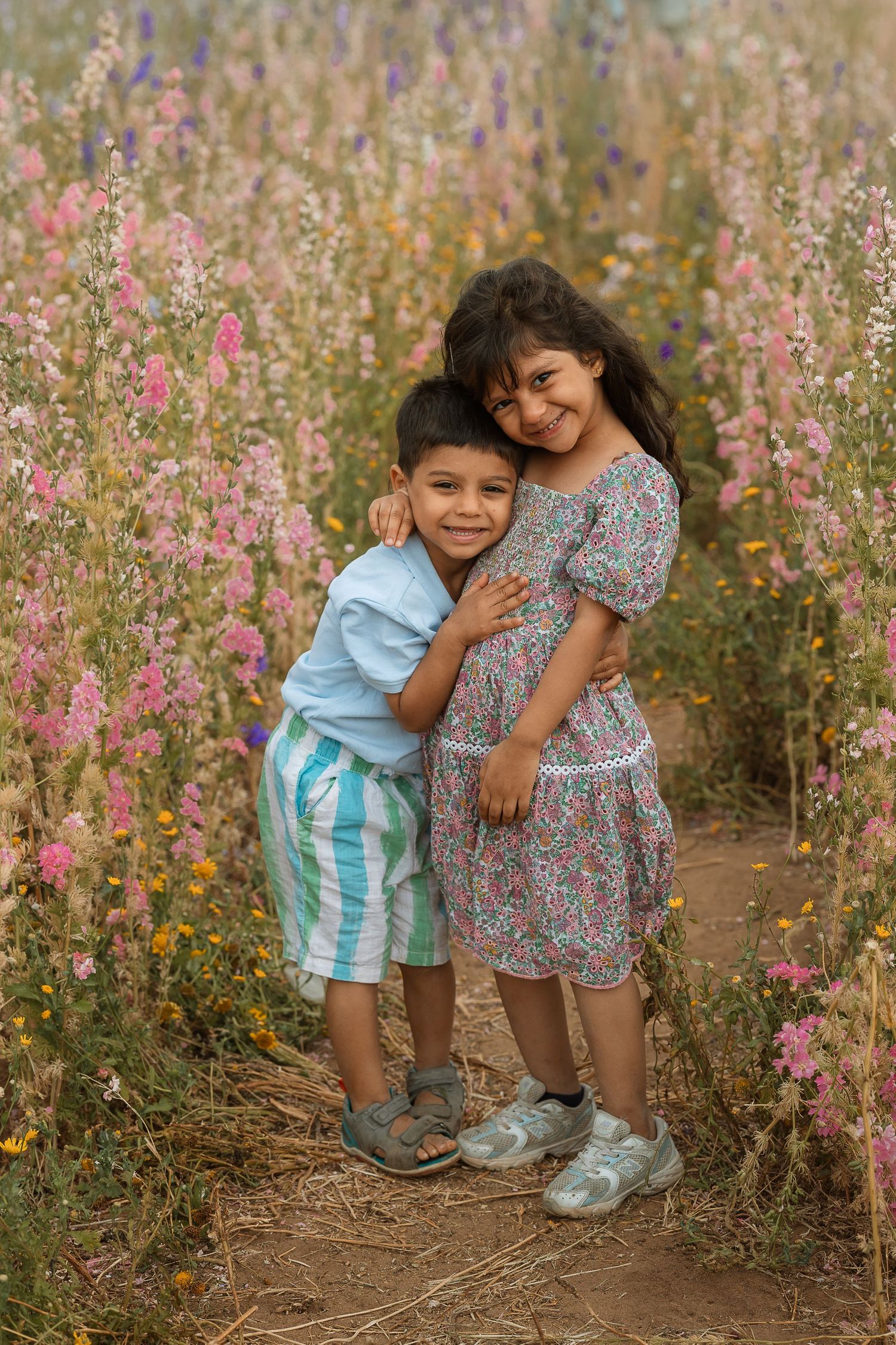 Two young children pose together among blooming pink wildflowers on a dirt path during golden hour.