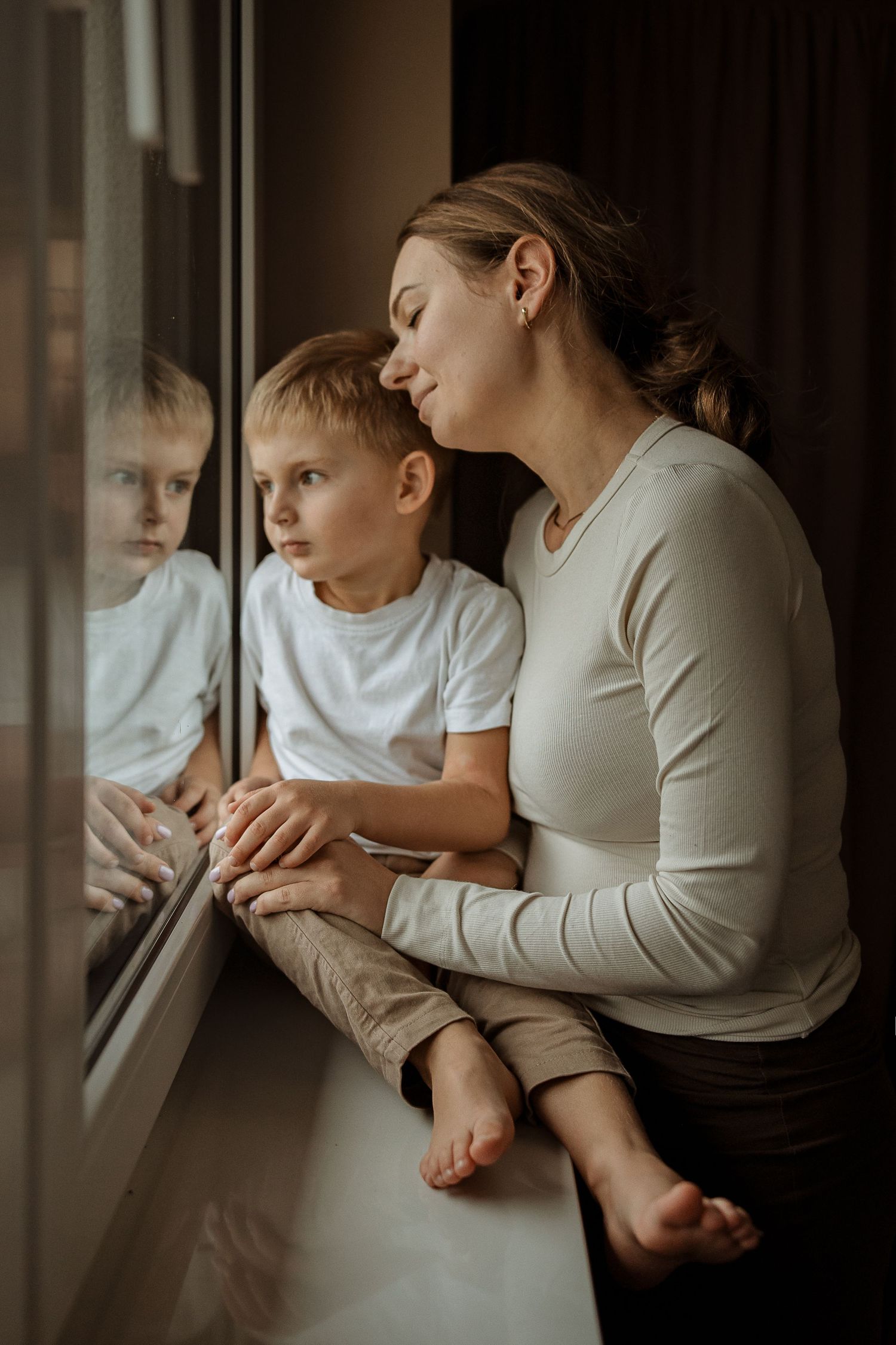 Mother and child sit by window in intimate black and white photo series showing tender moments together.