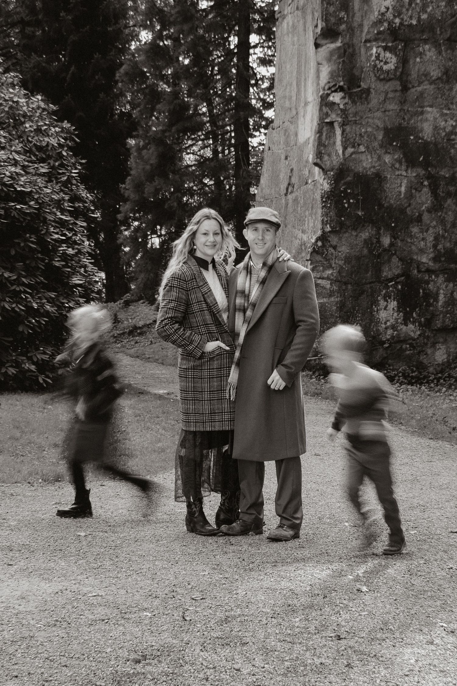 Black and white photo of a couple in winter coats standing outdoors while blurred figures run past in the background.