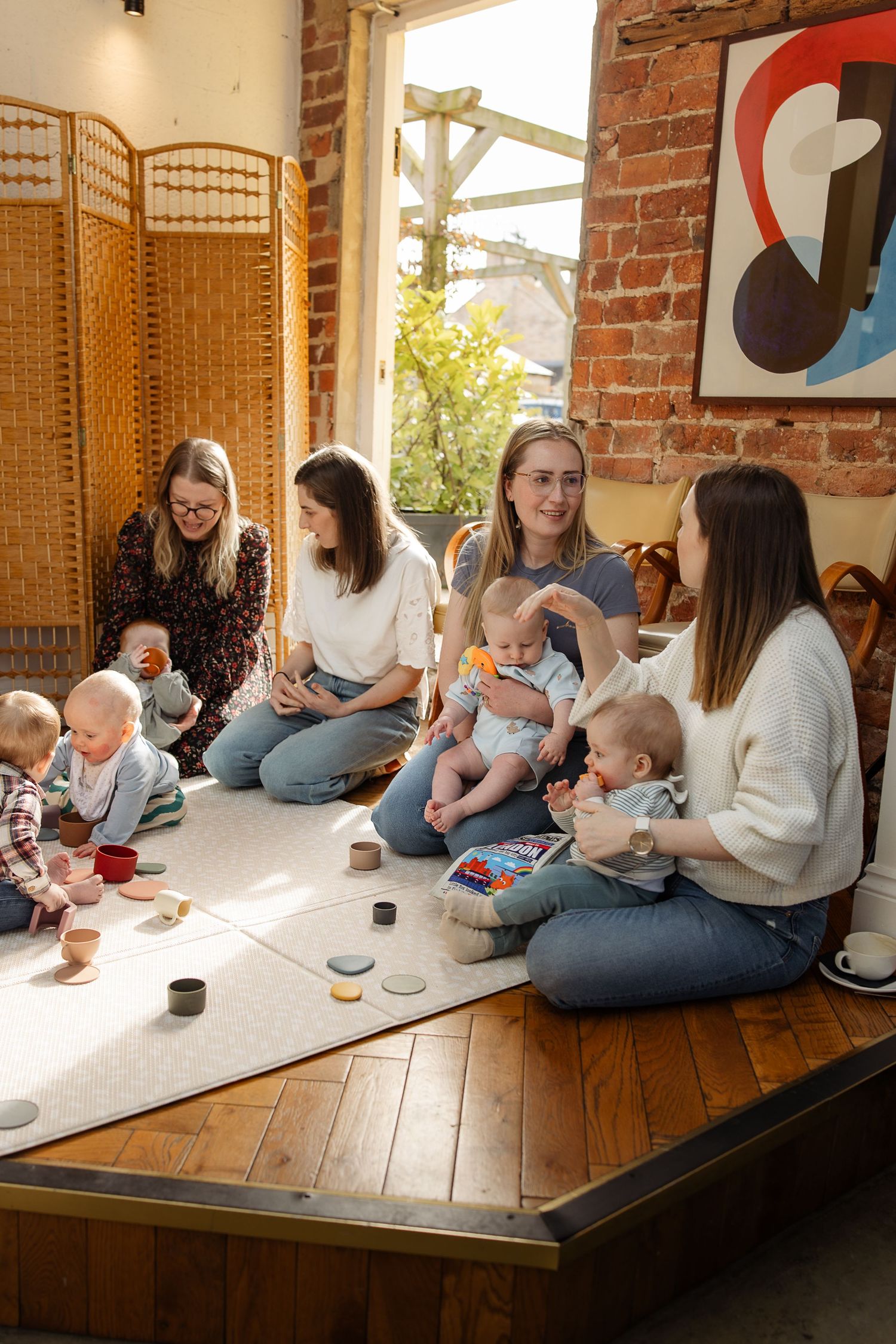 Parents and babies gather on a wooden floor against exposed brick walls during an indoor playgroup session.