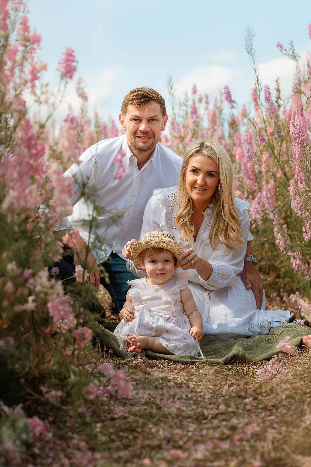 Happy family with baby girl wearing straw hat sits among pink wildflowers in a sunny field.