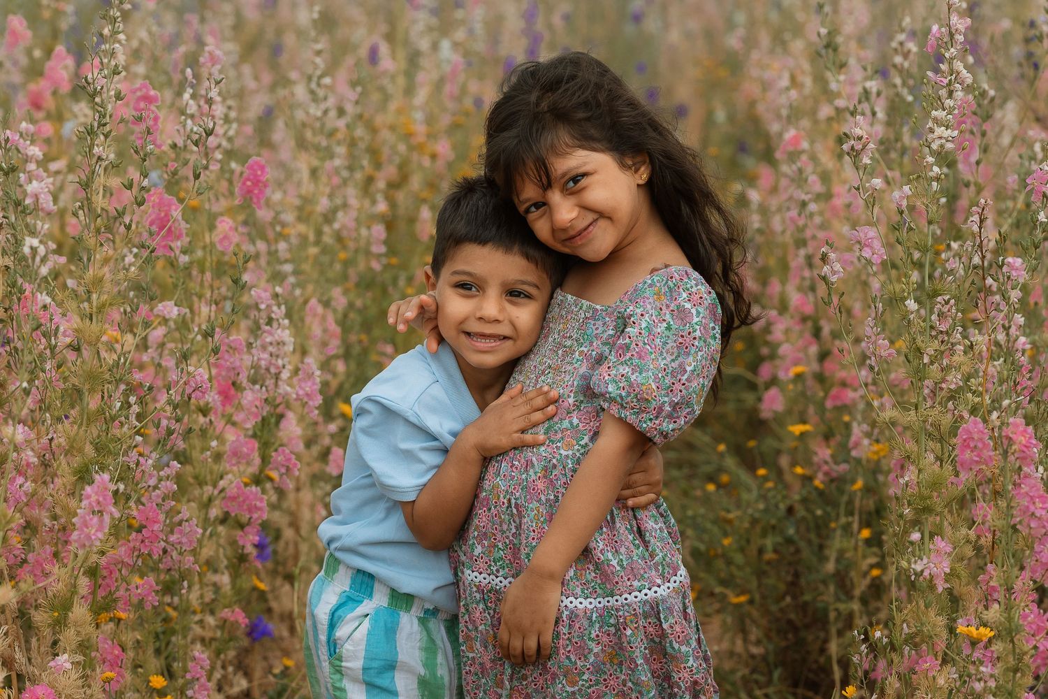 Two young siblings hugging in a vibrant wildflower field, smiling warmly among pink blooms.