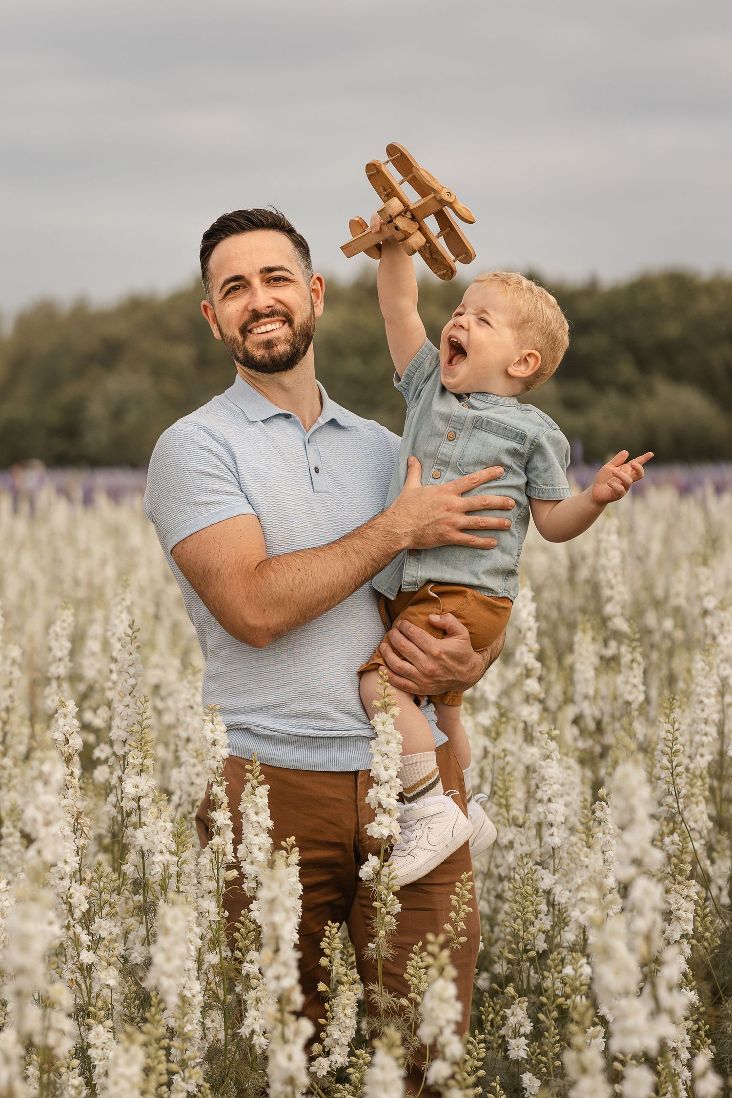 Father holding toddler playing with wooden toy airplane in white flower field, both smiling joyfully.