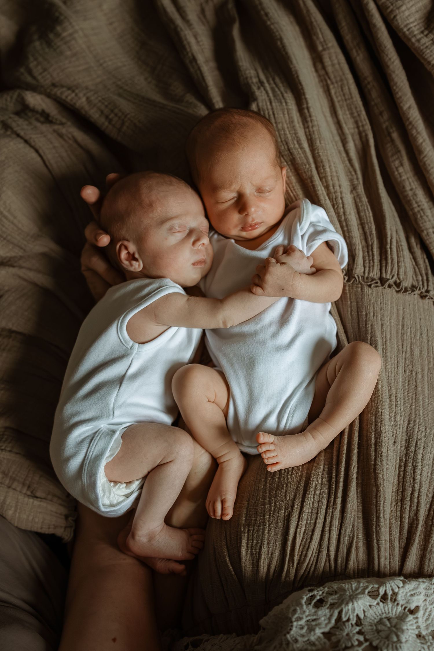 Two infants in white onesies lie peacefully together on a textured beige blanket in a series of intimate portraits.