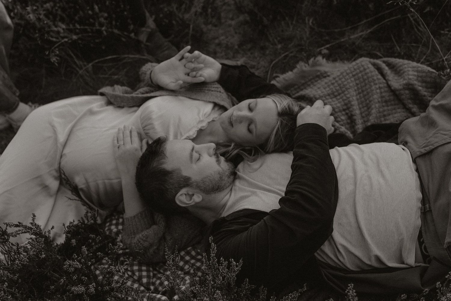 A black and white photograph shows two people lying together on grass in a tender moment outdoors.
