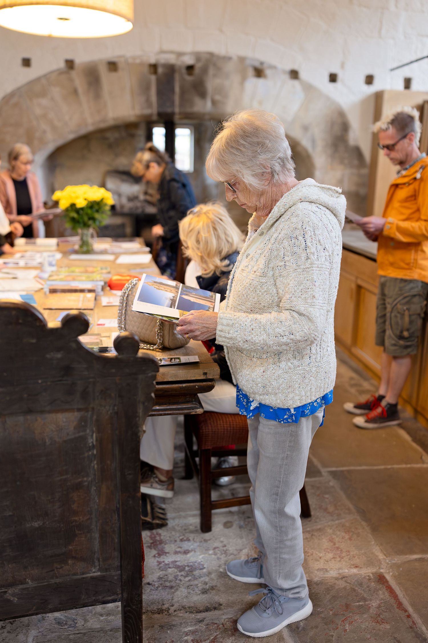 Elderly person in gray sweater and pants bending over to look at items in a rustic stone-walled space.