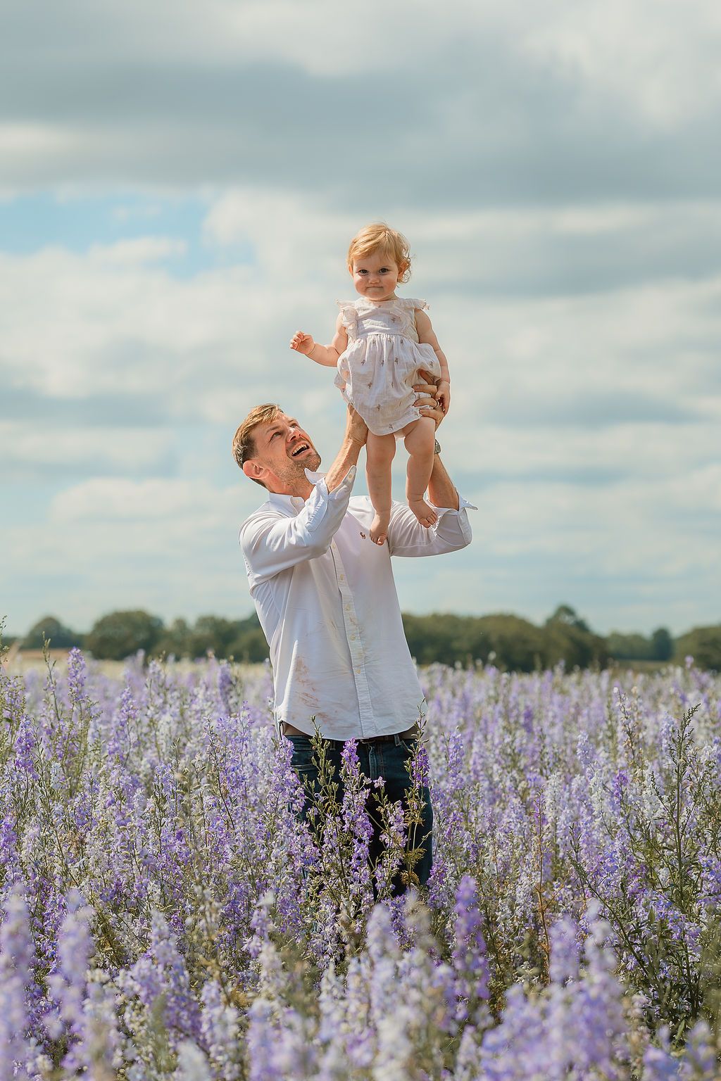 Father joyfully lifting baby girl in purple flower field under cloudy sky.