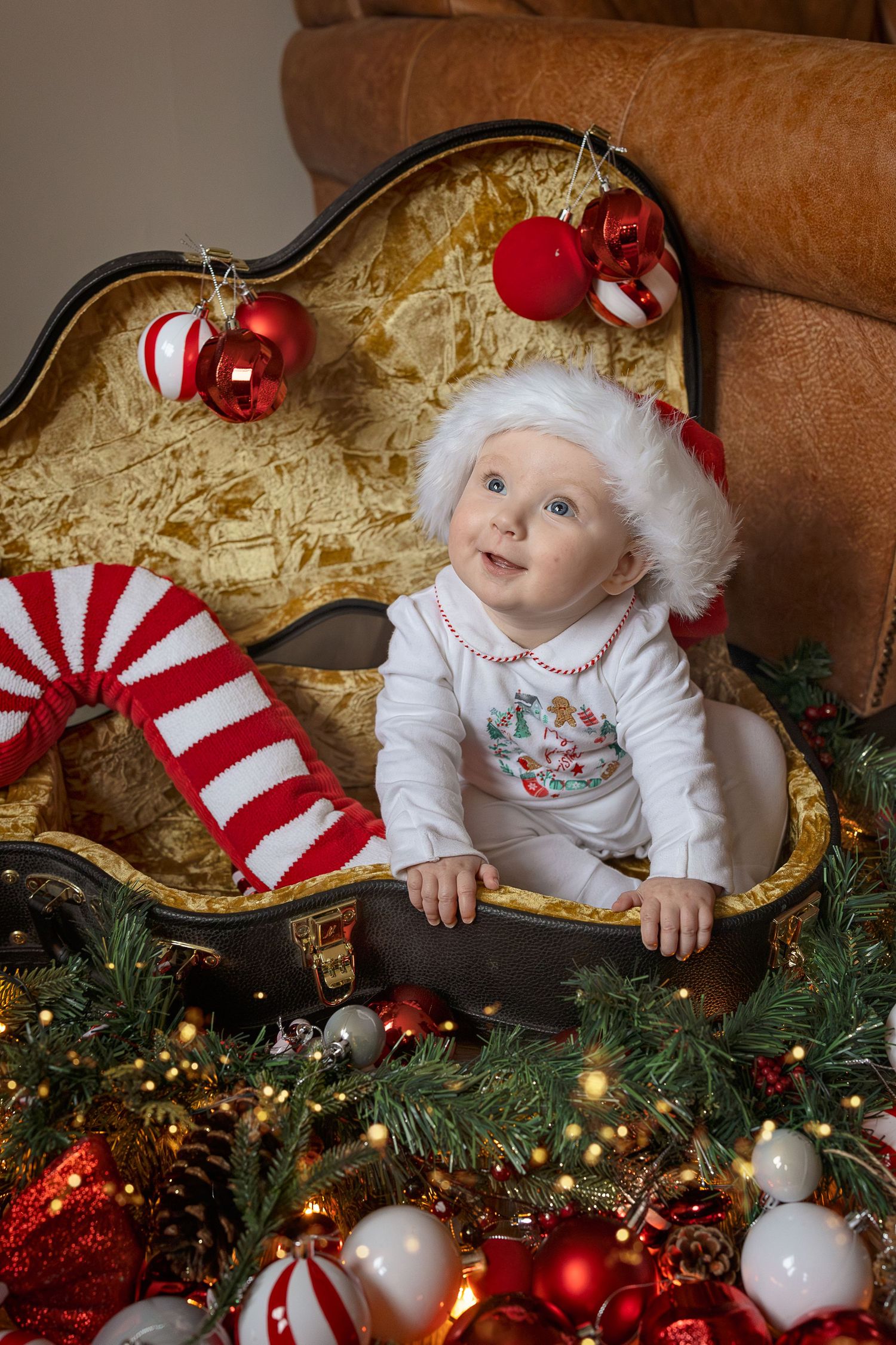 Baby in Santa hat sits in vintage suitcase surrounded by Christmas ornaments, candy canes, and festive decorations.