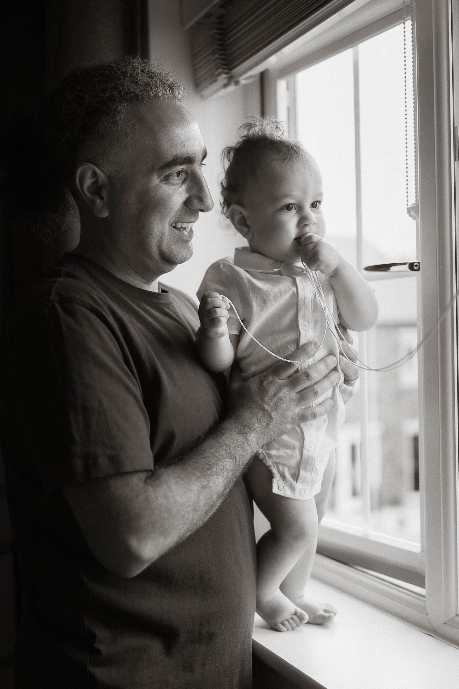 A tender black and white photo series showing an adult and baby looking out a bright window together.