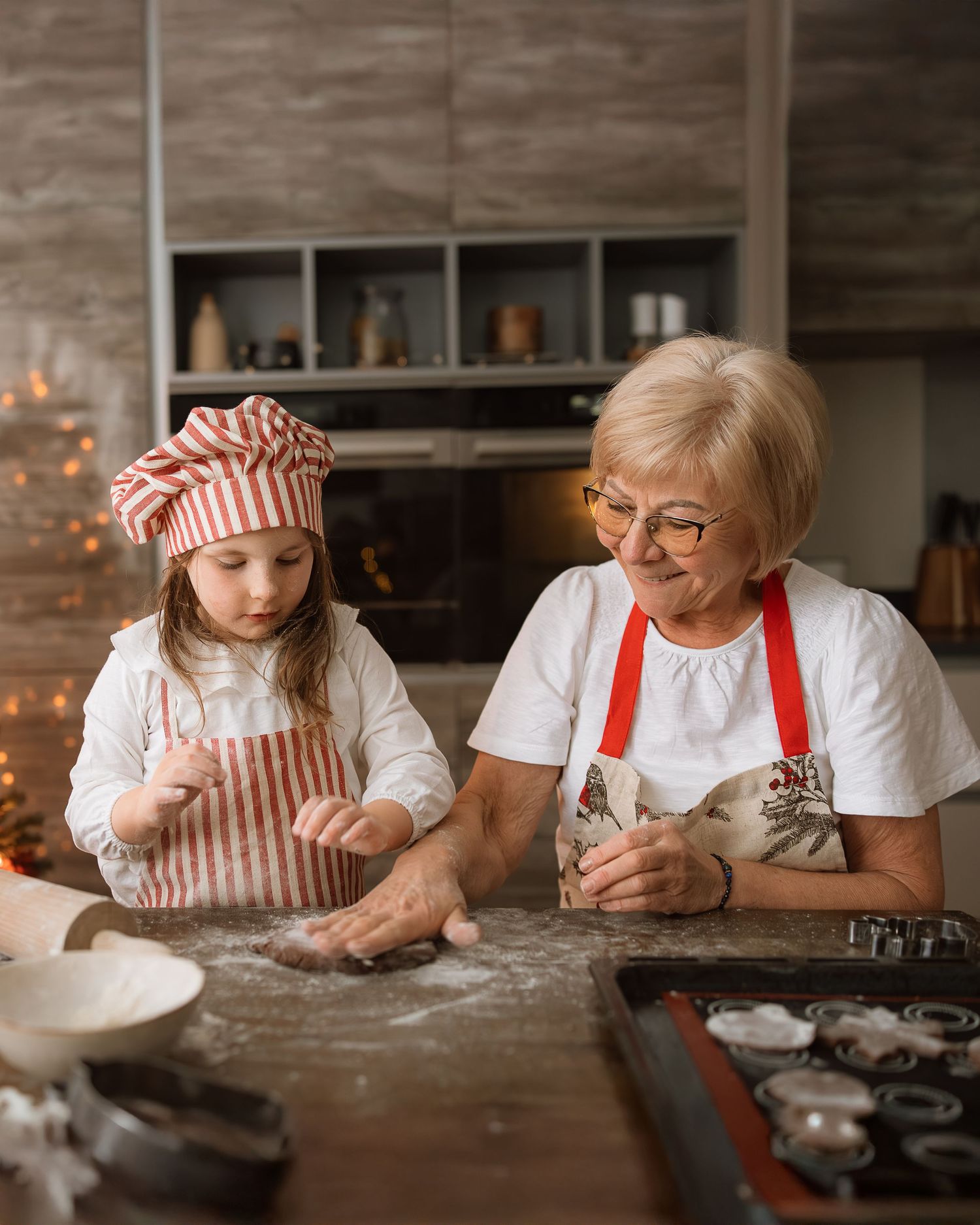 Grandmother and granddaughter baking Christmas cookies together in a cozy kitchen, wearing matching red aprons and chef hats.