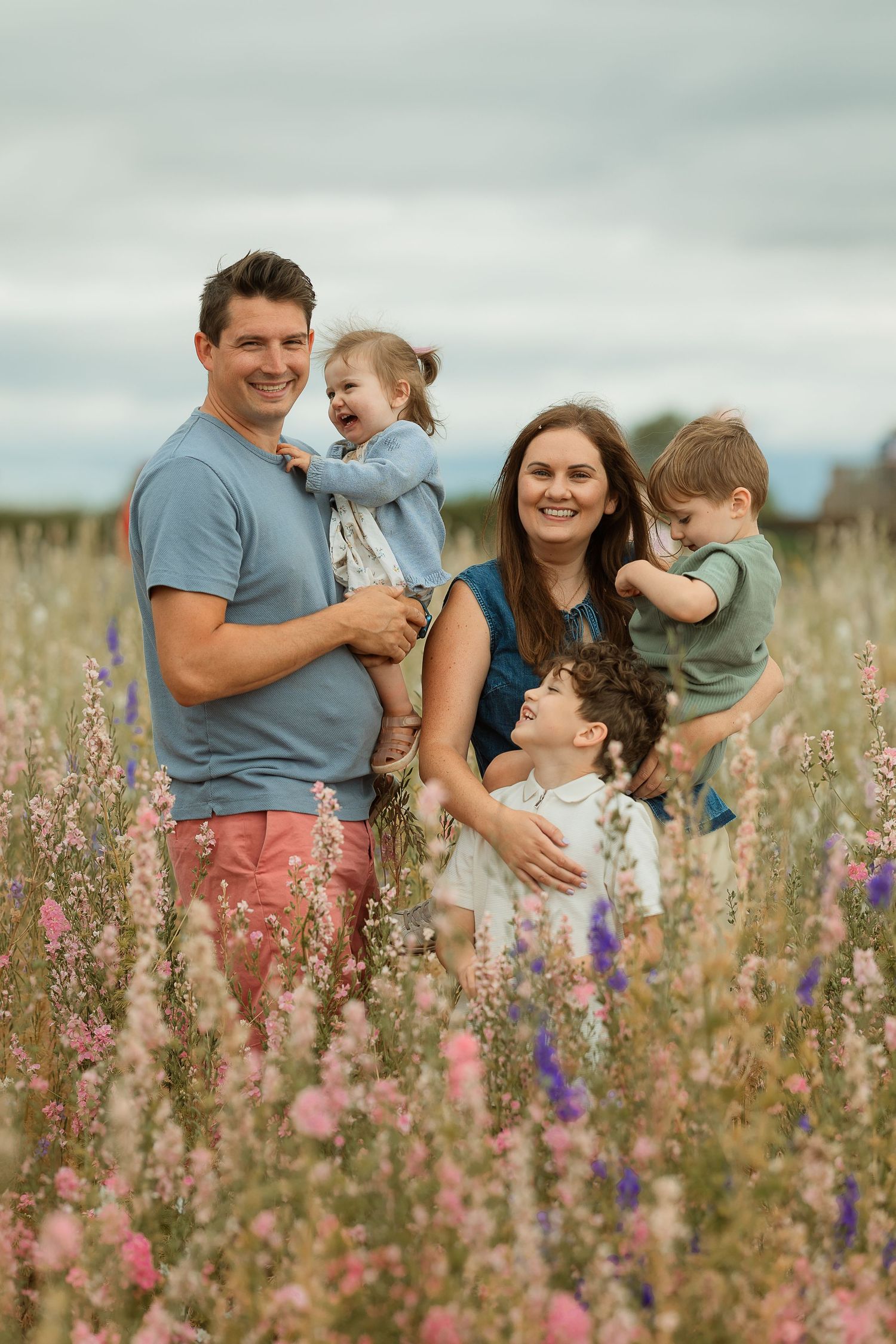 A family shares joyful moments together in a wildflower meadow filled with pink and purple blooms on a cloudy day.