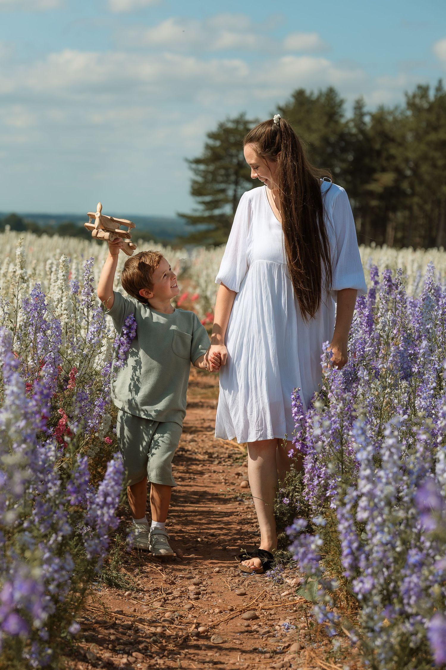 People walking through rows of blooming purple delphinium flowers on a sunny summer day in a garden field.