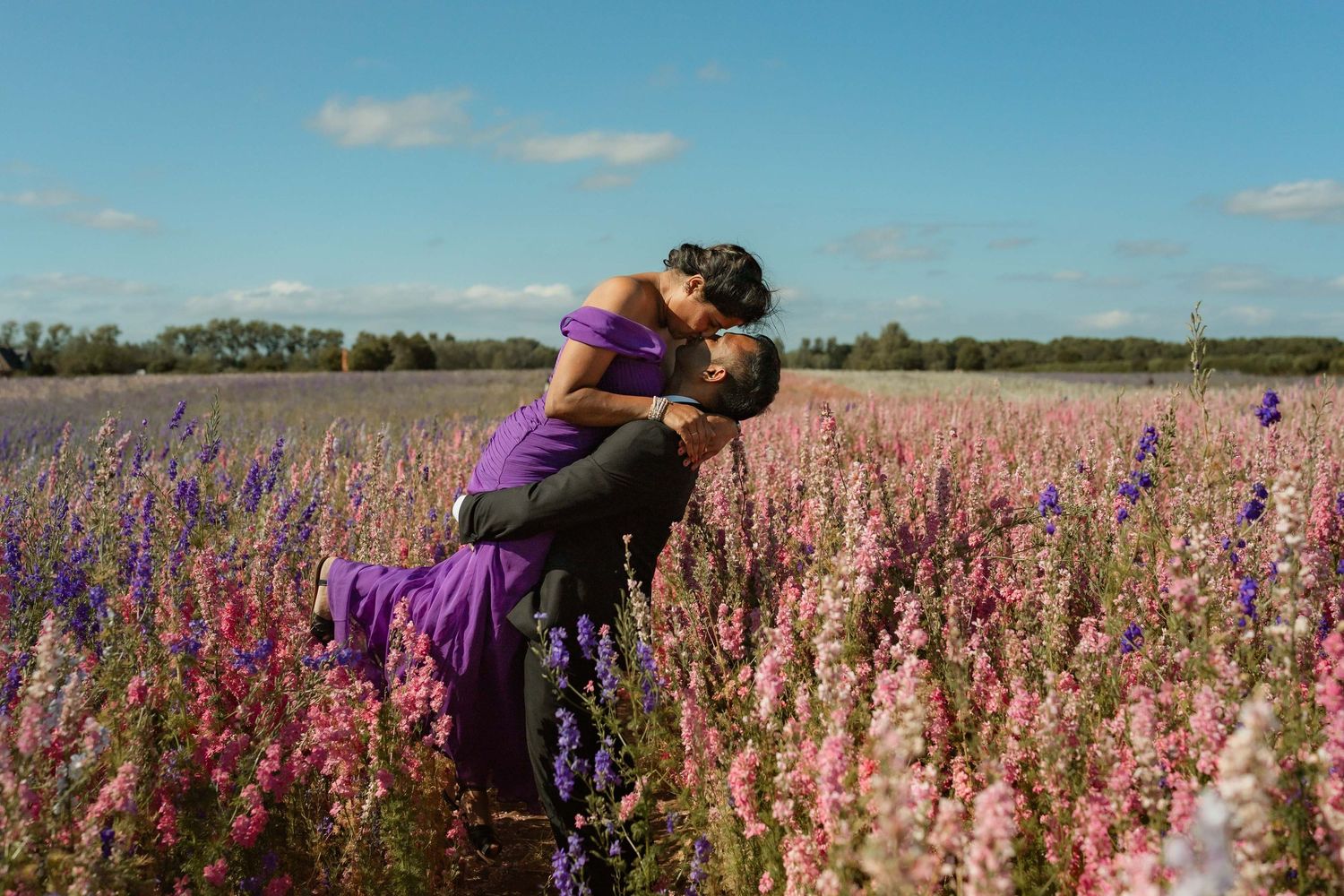 Couple kissing in a vibrant field of pink and purple wildflowers under a bright blue sky.