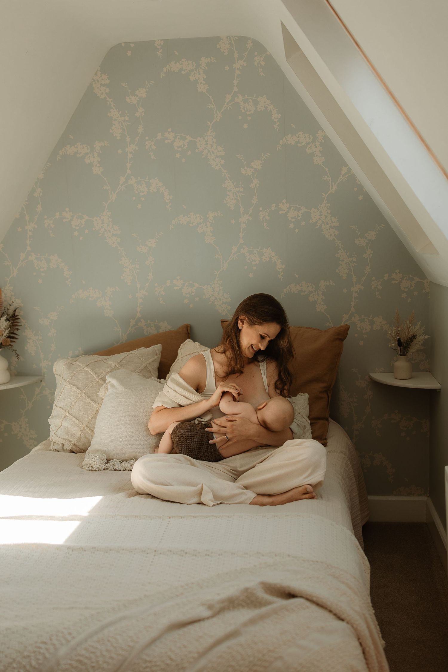 A person rests peacefully on a bed with floral wallpaper and soft white bedding in natural sunlight.