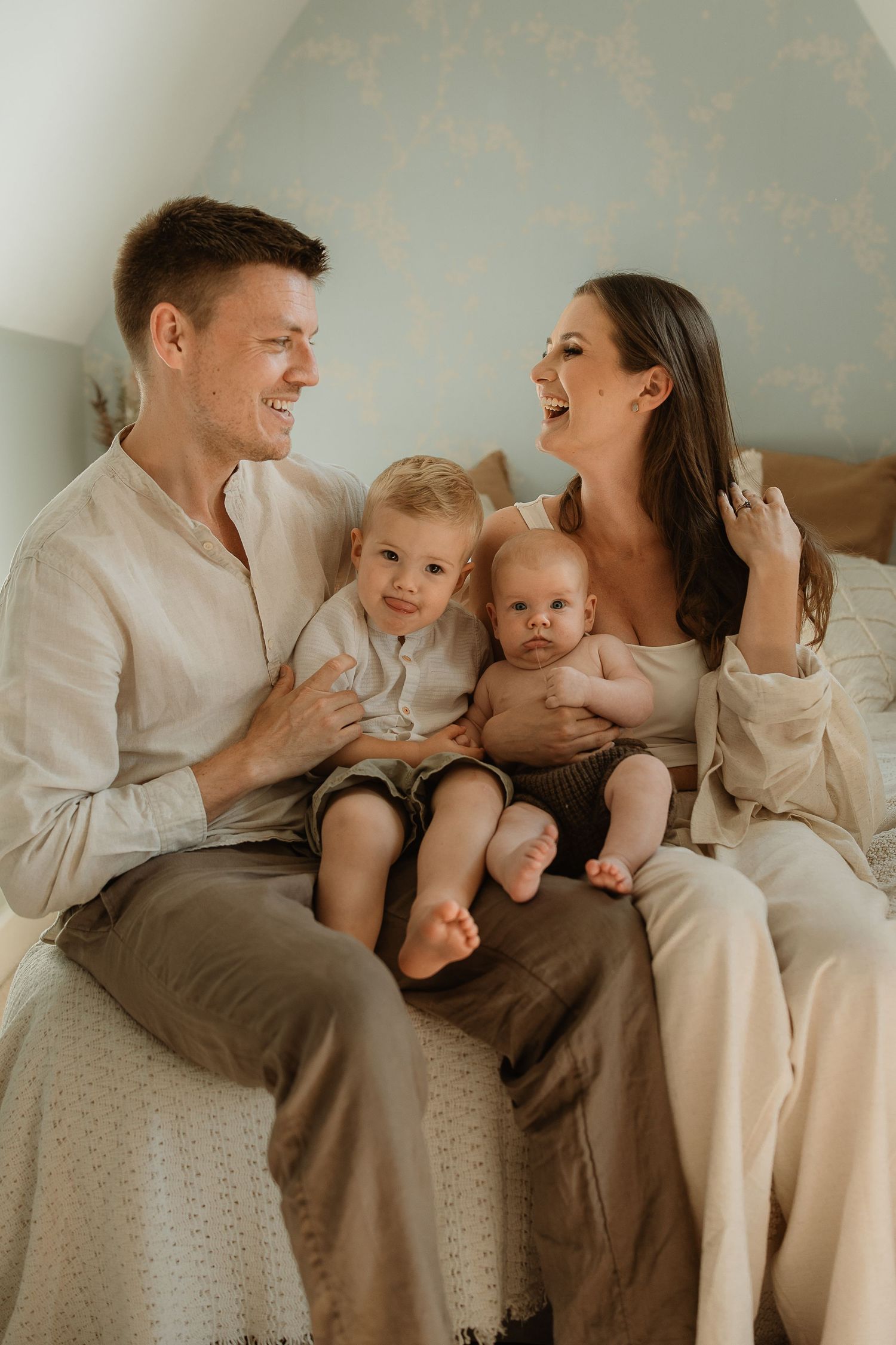 A joyful family moment captured on a bed with two young children in casual neutral-toned clothing.