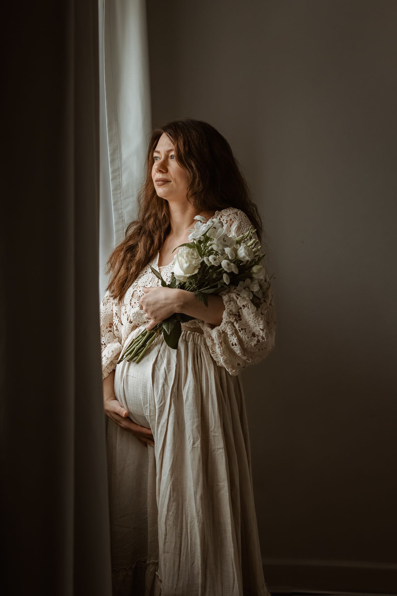 Moody boudoir portrait series of woman in white nightgown holding white flowers by window in dark room.