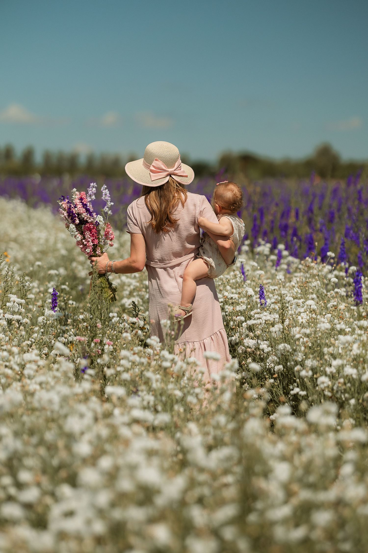 A woman in a pink dress and sun hat holds a baby while carrying wildflowers in a blooming field of white and purple flowers.