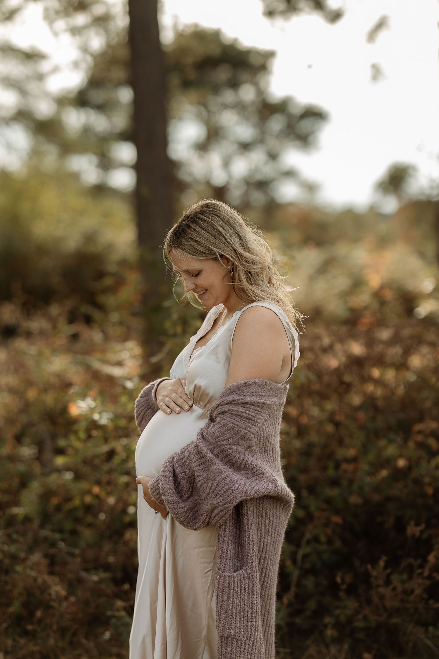 A person in a cozy knit sweater embraces their pregnant belly in a serene outdoor setting with autumn foliage.