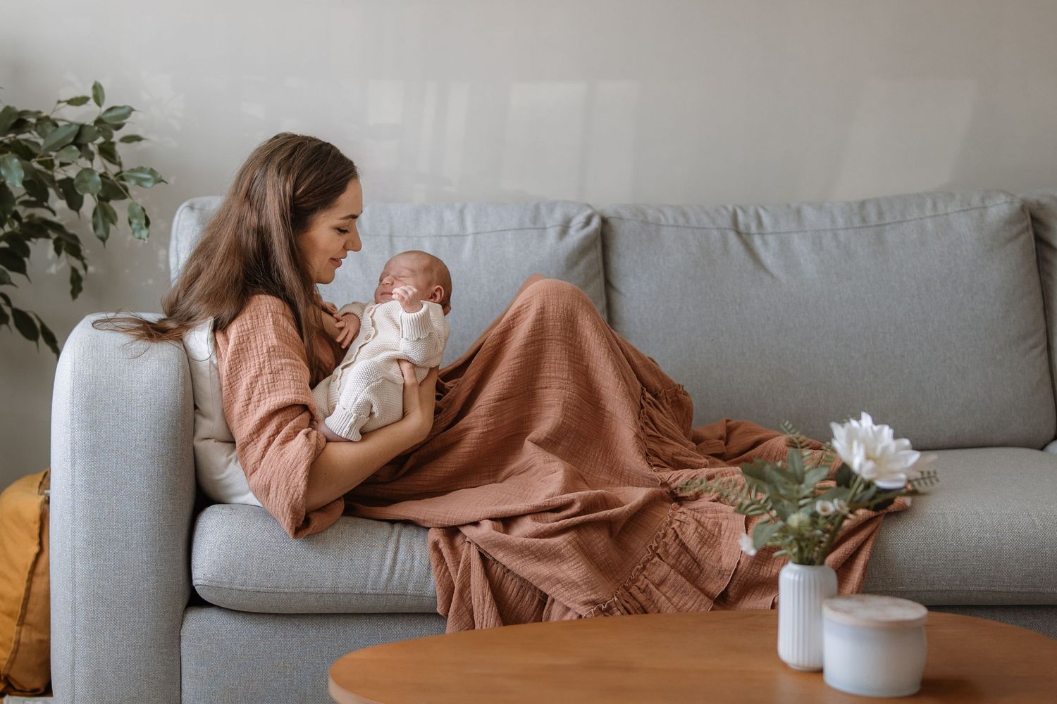 A person relaxes on a gray couch in a cozy living room with warm brown blankets and indoor plants.