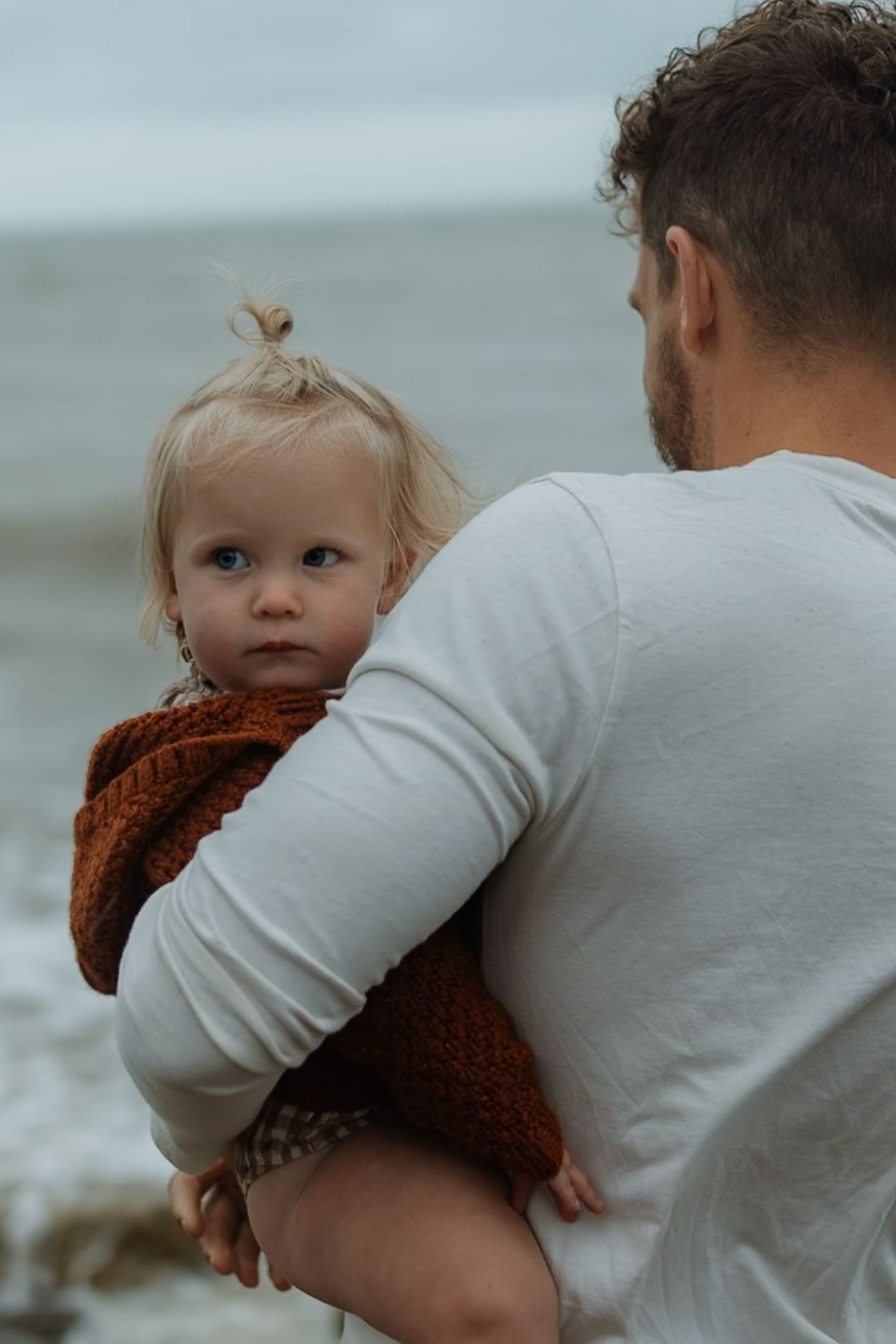 A parent in a white shirt holds a small child wearing a brown sweater against an overcast coastal background.