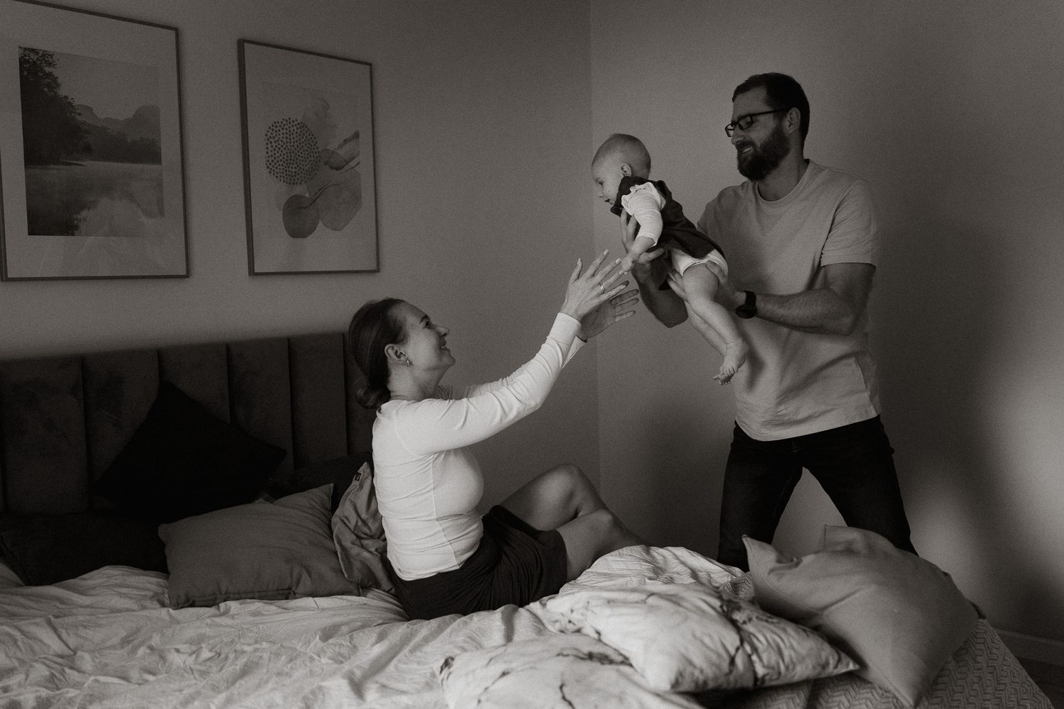Parents play with their baby on a bed in a black and white photograph with framed artwork on the wall.