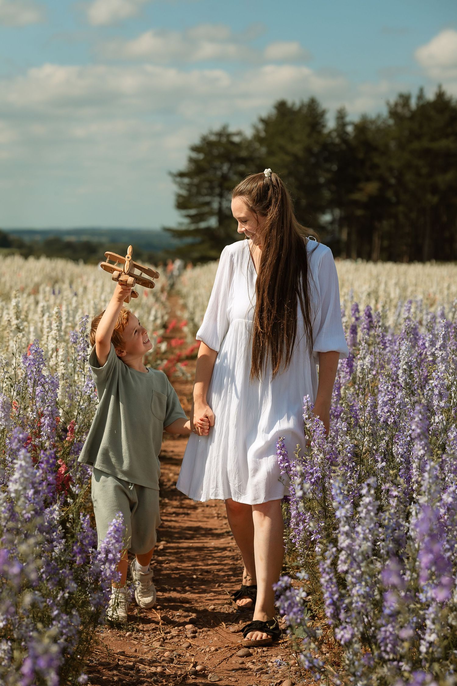 A parent and child walk together on a dirt path through a beautiful field of purple wildflowers on a sunny summer day.