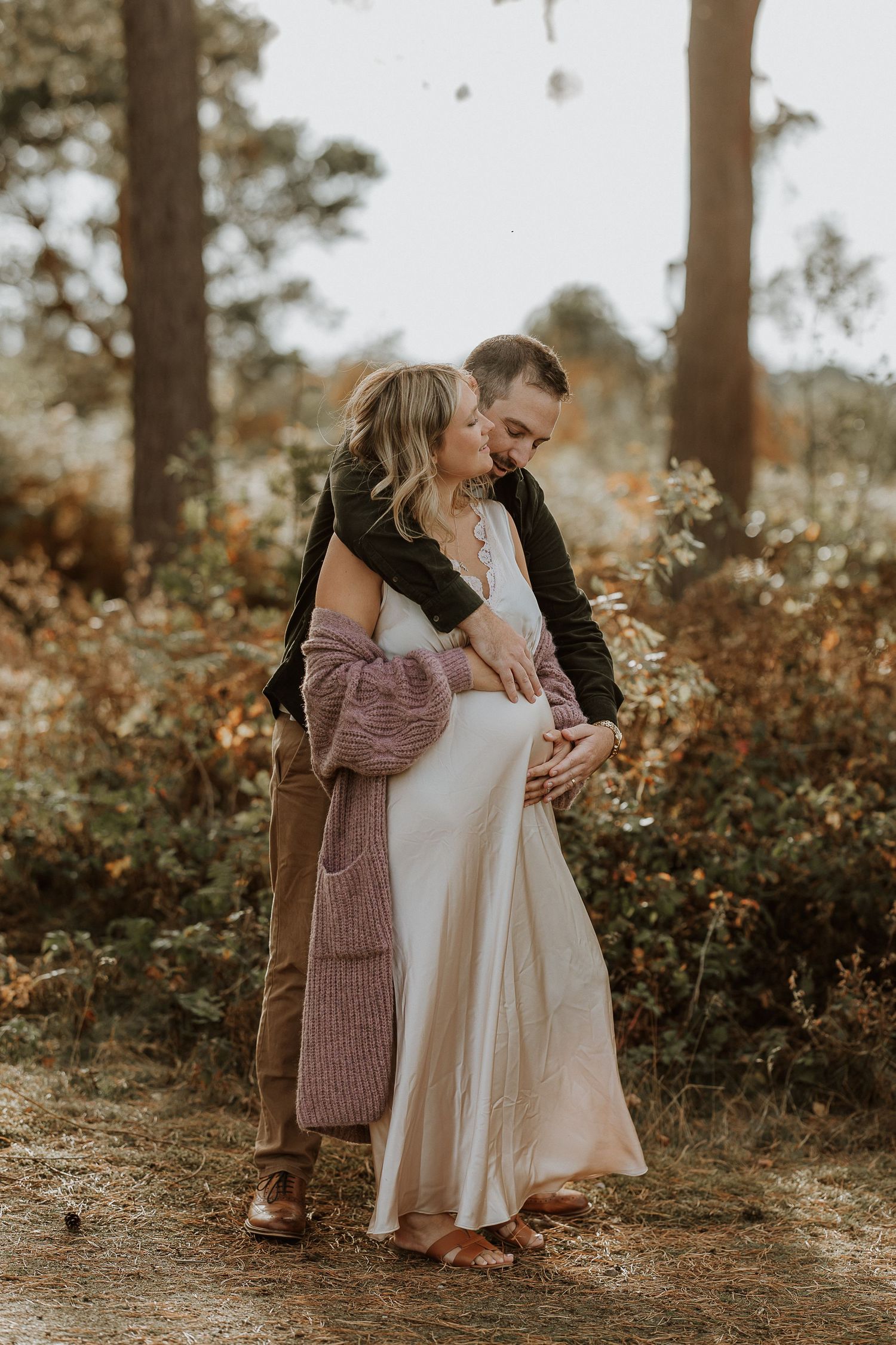 Family embraces in a sunlit forest setting wearing cozy fall outfits during an outdoor photo session.
