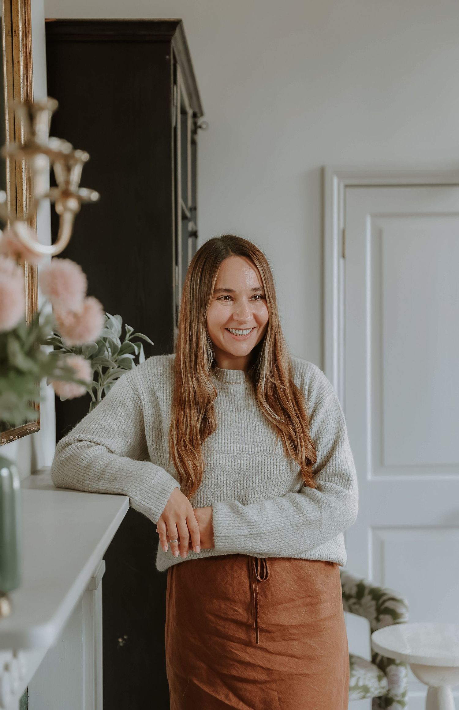 Person wearing a cozy beige sweater and brown skirt poses playfully in a bright interior space.