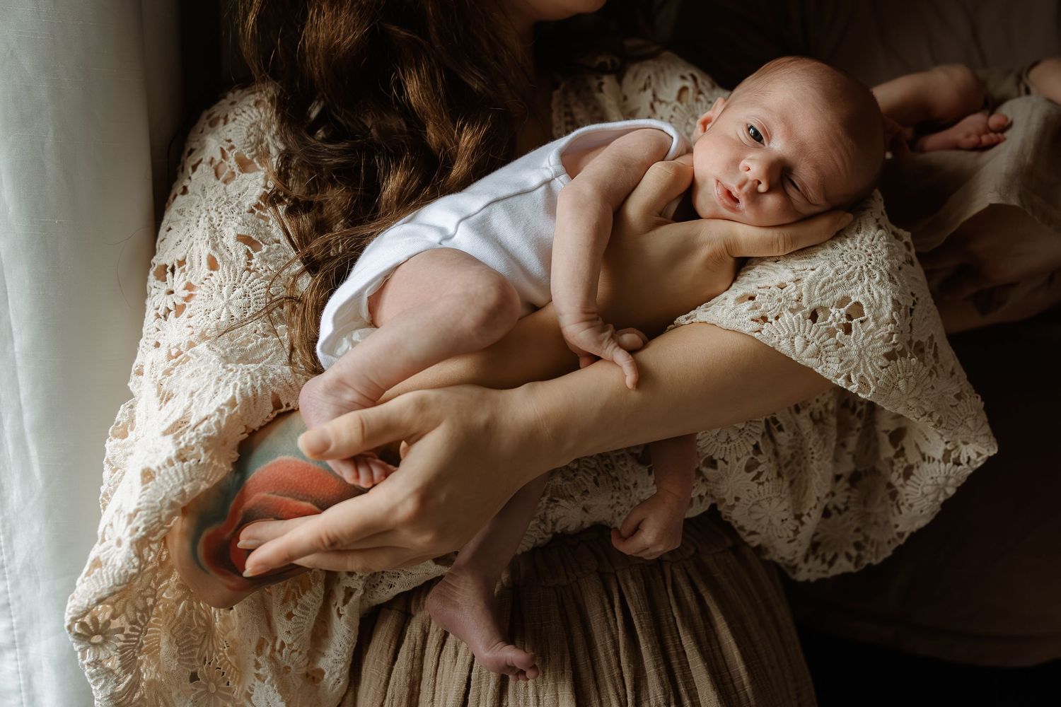 A small baby in a white onesie rests peacefully in loving arms on a lace-covered chair.