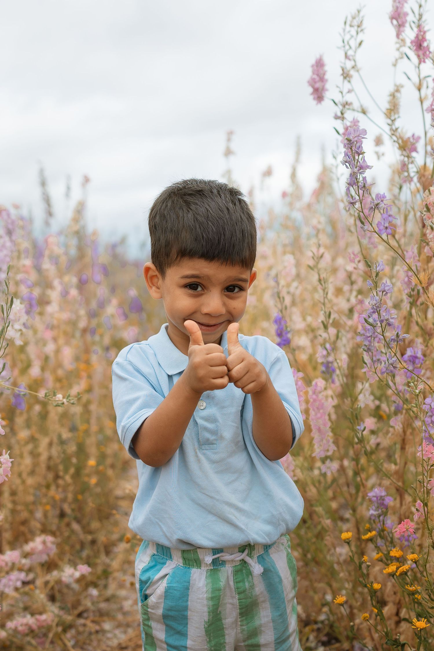 Young boy giving thumbs up in a colorful wildflower field, smiling cheerfully outdoors.