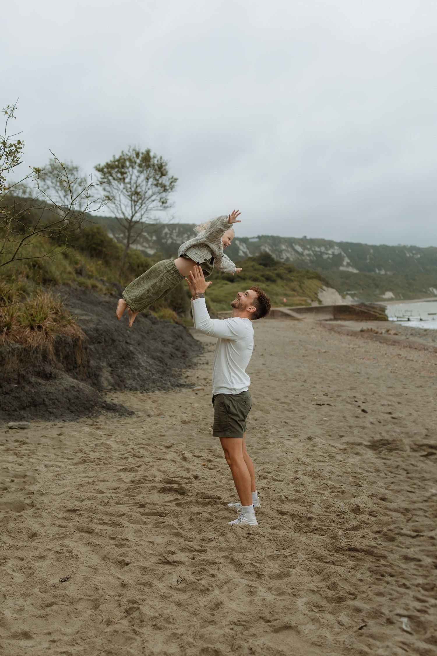 A person in casual attire performs a series of jumping poses on a misty beach with hills in the background.