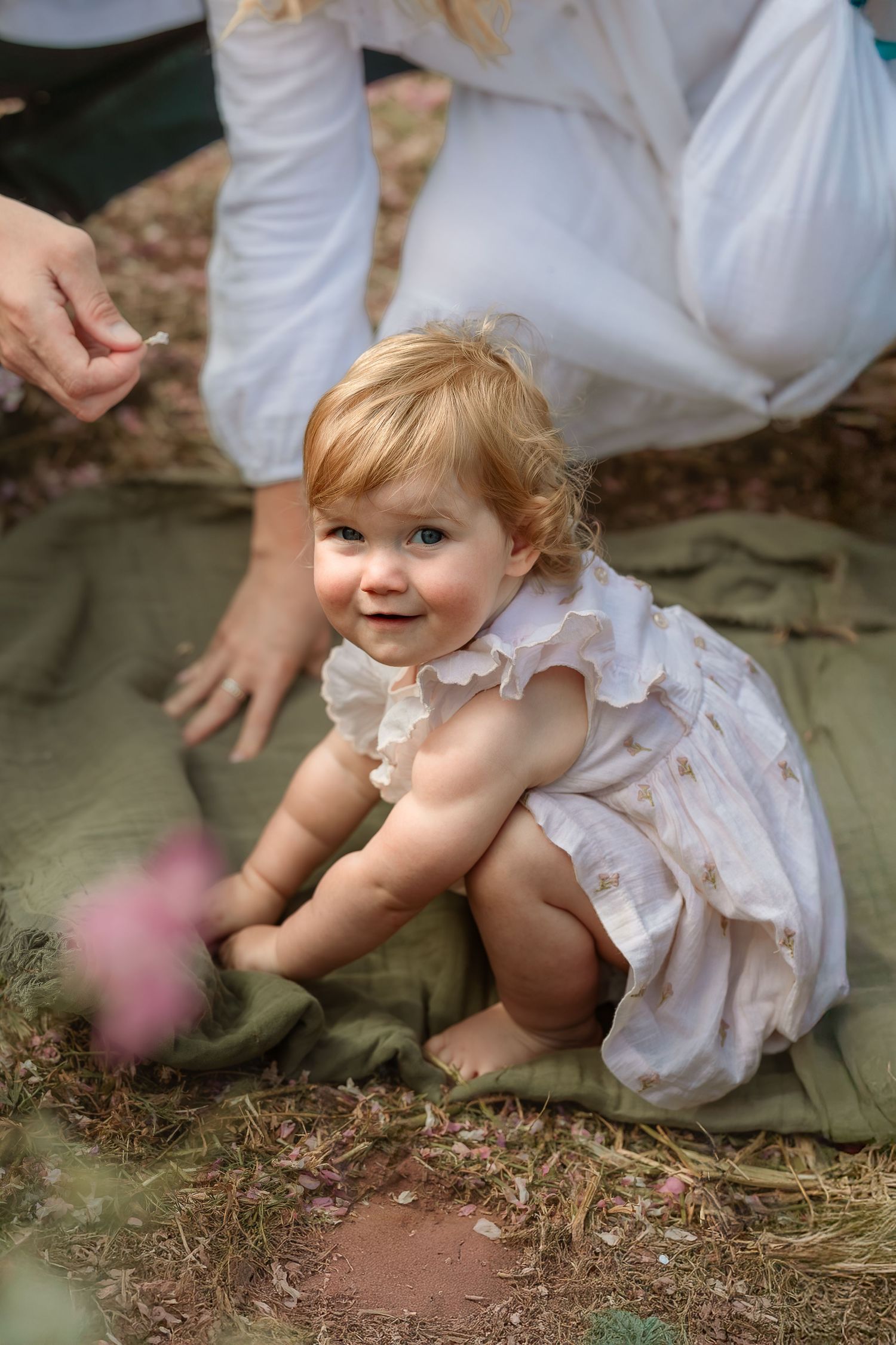 A baby in a white dress sits playfully on the ground in a series of candid outdoor photos.
