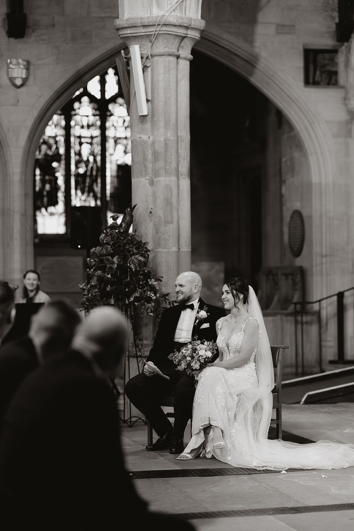 A black and white wedding ceremony photo showing a couple seated under a grand stone archway in a church.