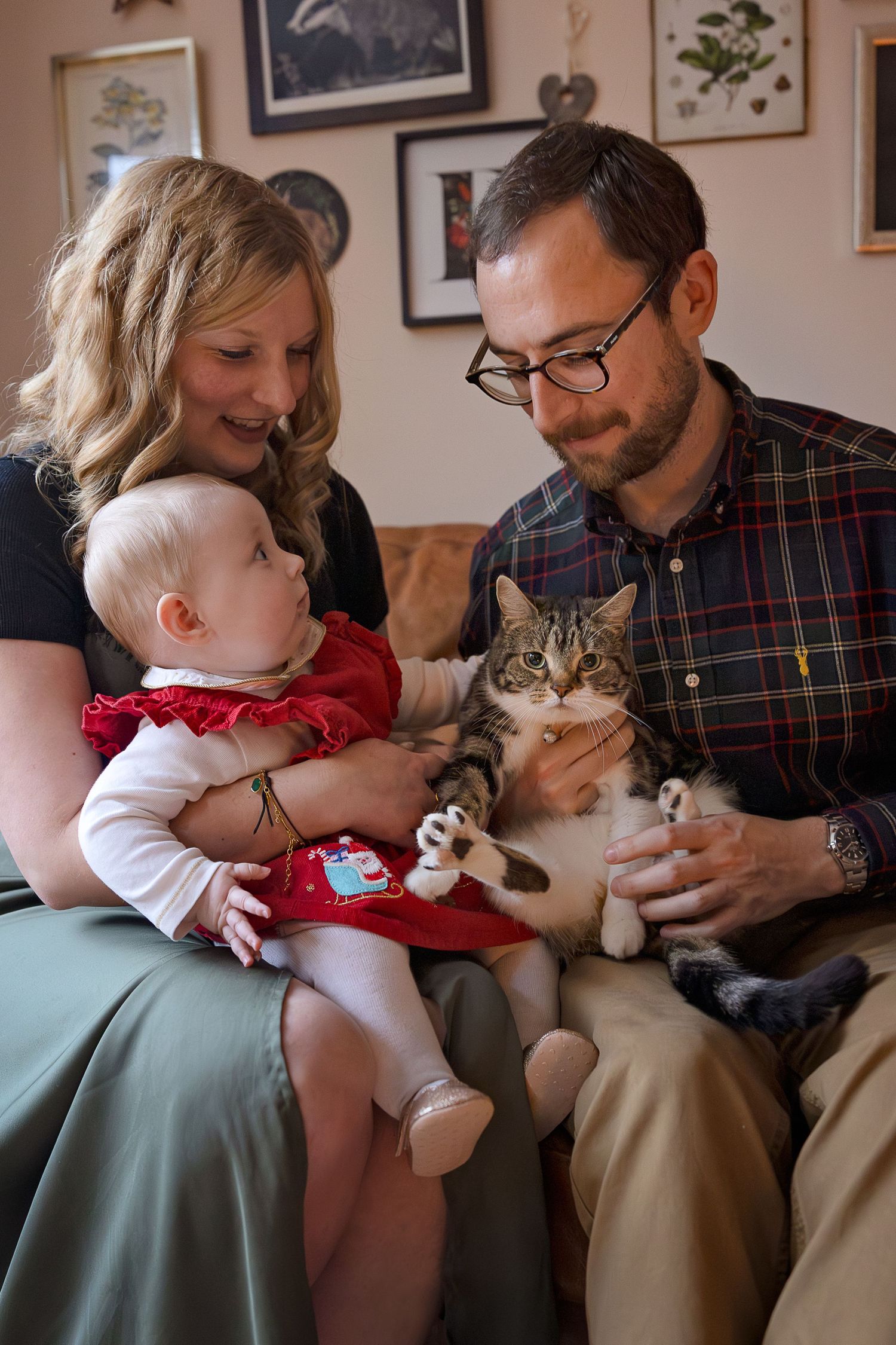A happy family with a baby in red dress and a tabby cat sitting together on a couch at home.