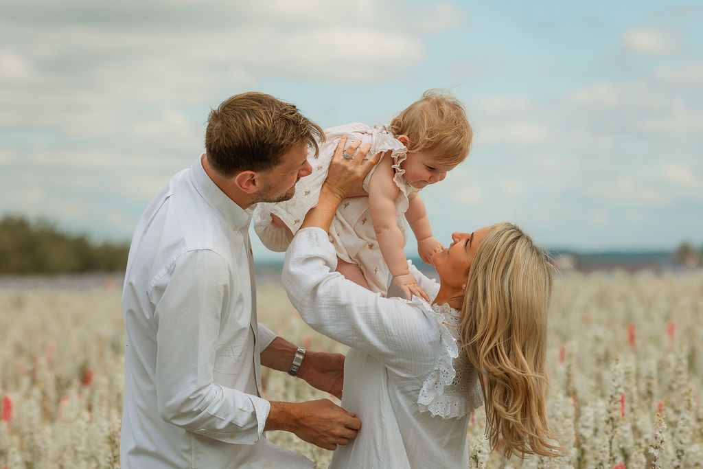 Happy family with parents lifting smiling baby girl in a blooming field under a cloudy sky.