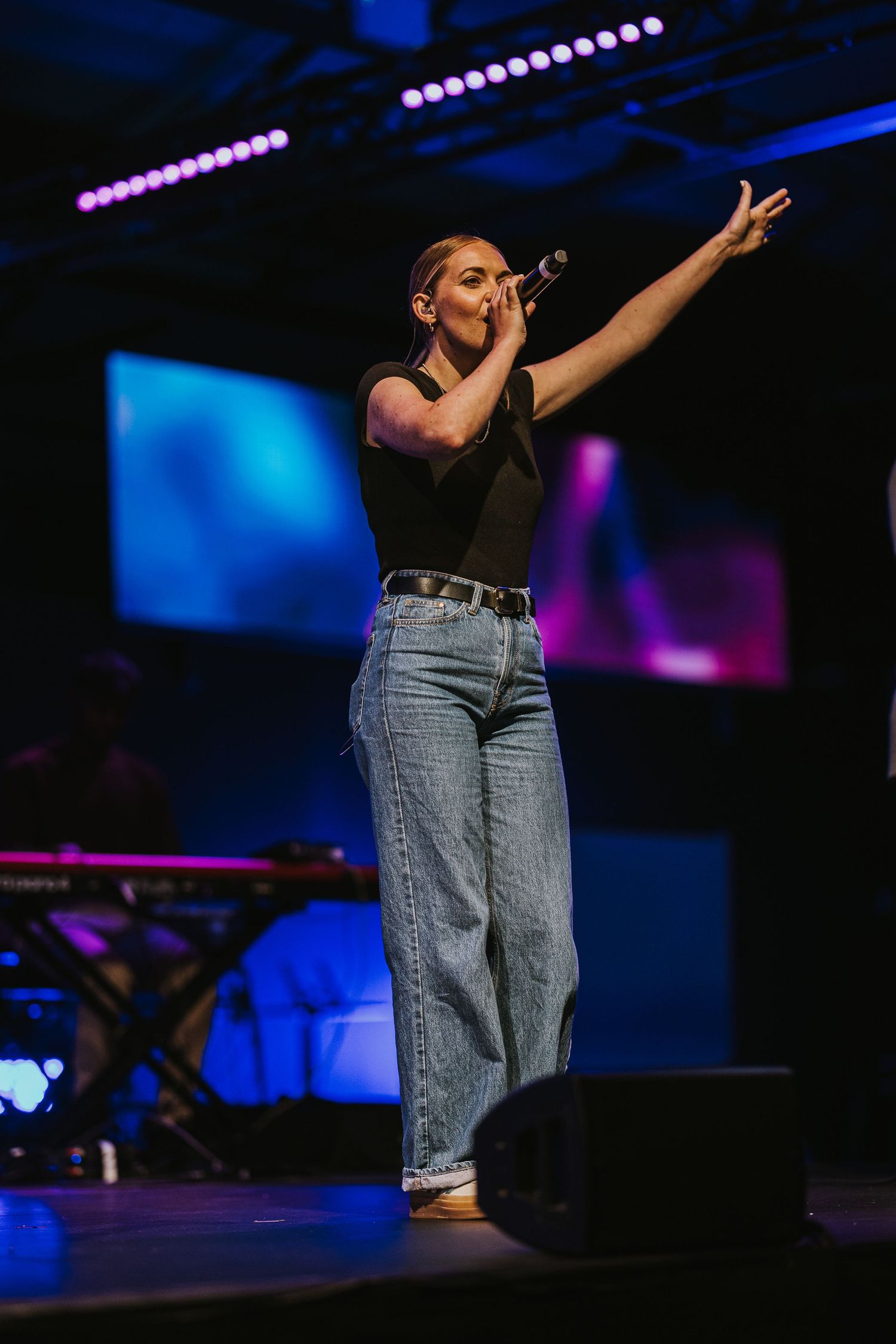 A performer in jeans sings passionately on stage with dramatic lighting and blue backdrop during a live show.