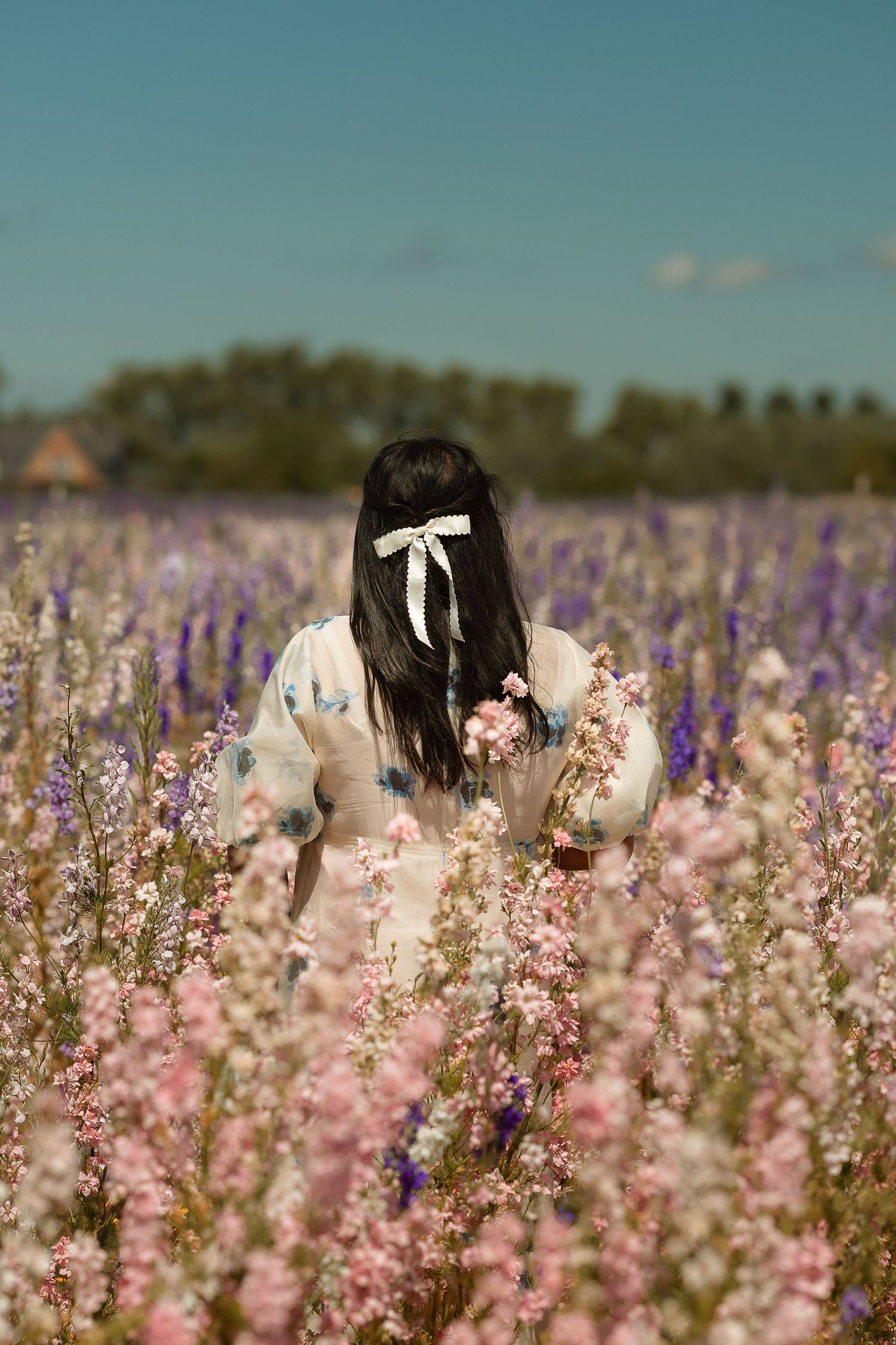 Young woman with dark hair and white bow standing in a blooming field of pink and purple wildflowers.