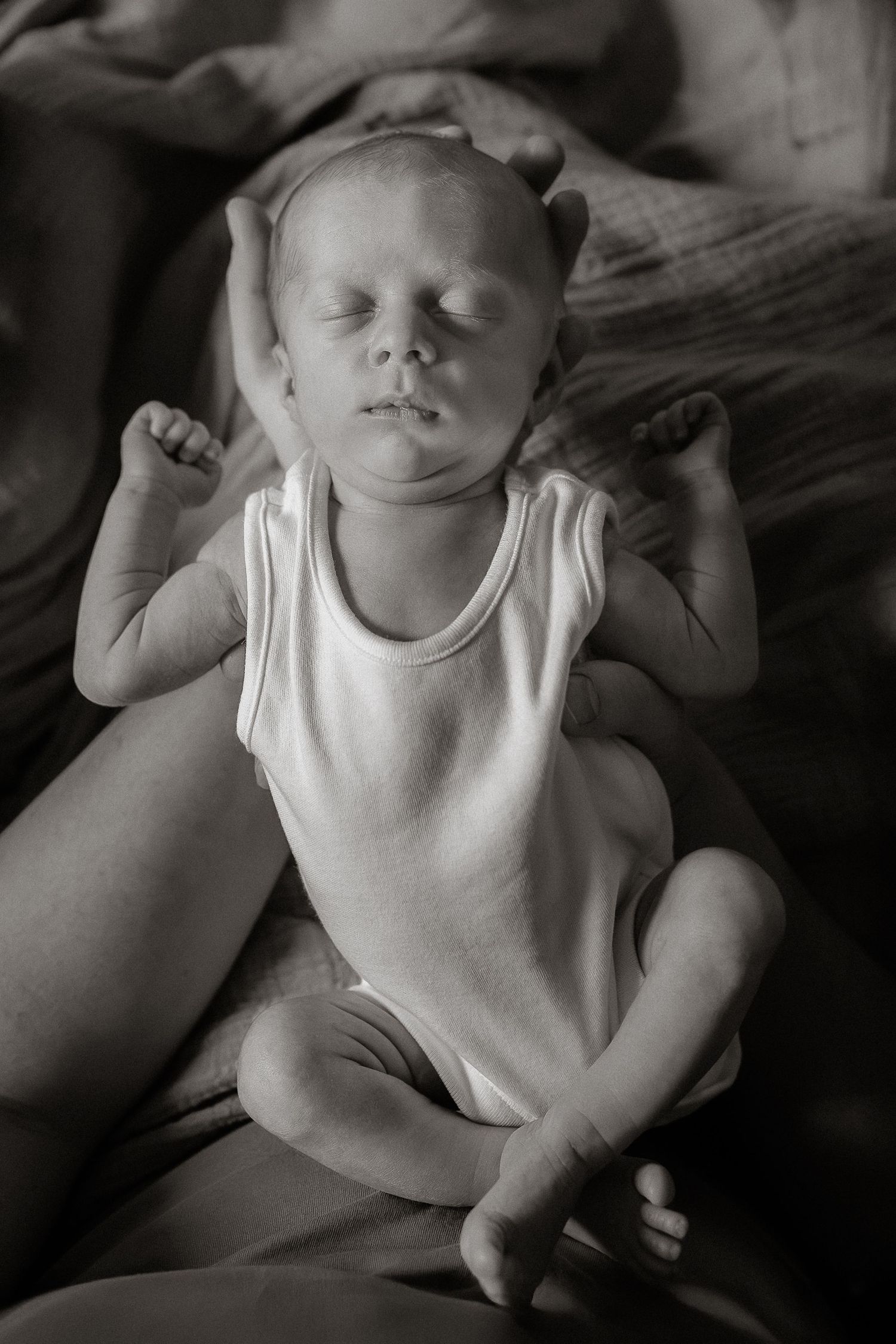 A series of black and white photographs showing a sleeping baby in a white sleeveless outfit on dark fabric.
