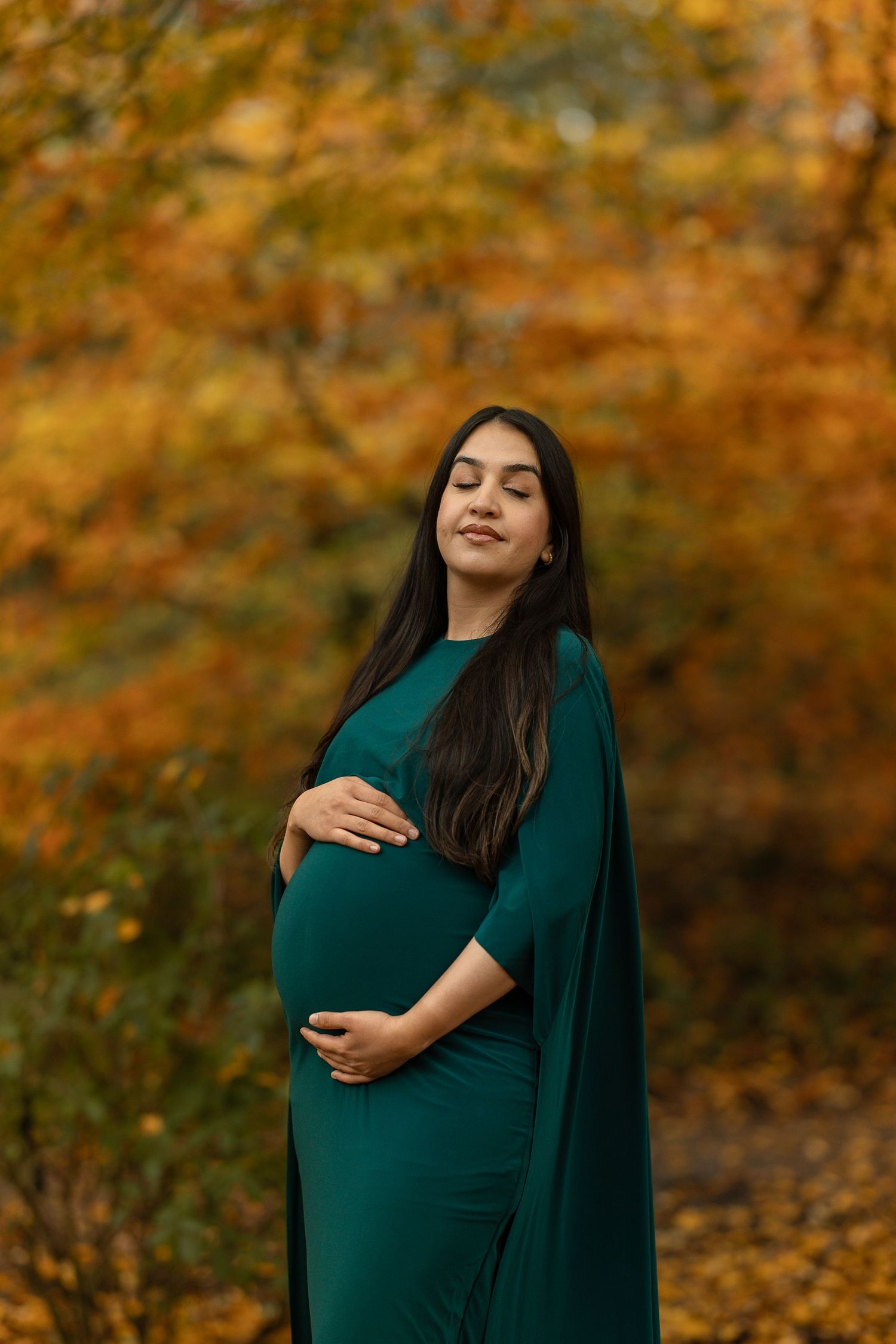 A person in a dark green dress poses gracefully against a backdrop of golden autumn foliage.