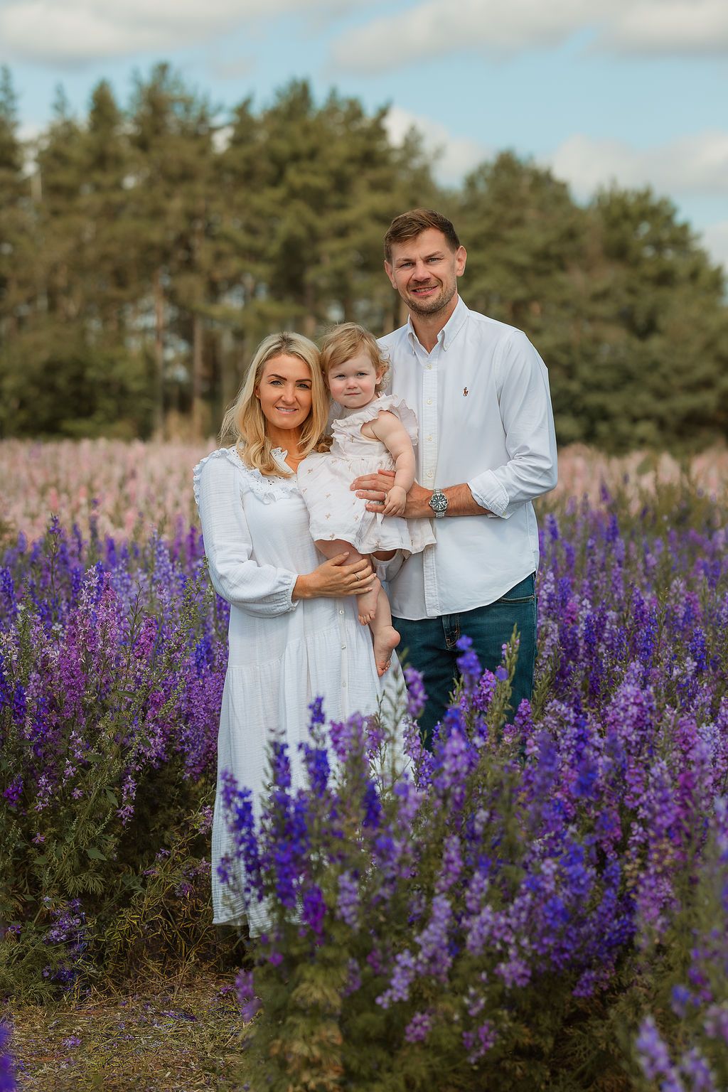 A happy family of three poses in a vibrant purple flower field, with tall green trees in the background.