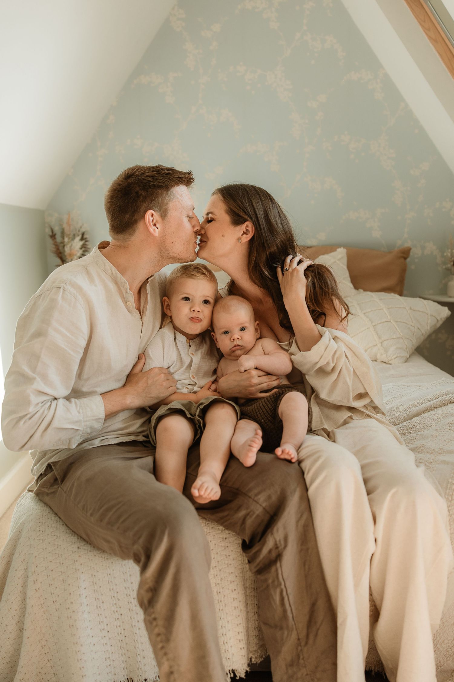 A family shares tender moments together while sitting on a bed in matching white and neutral toned outfits.