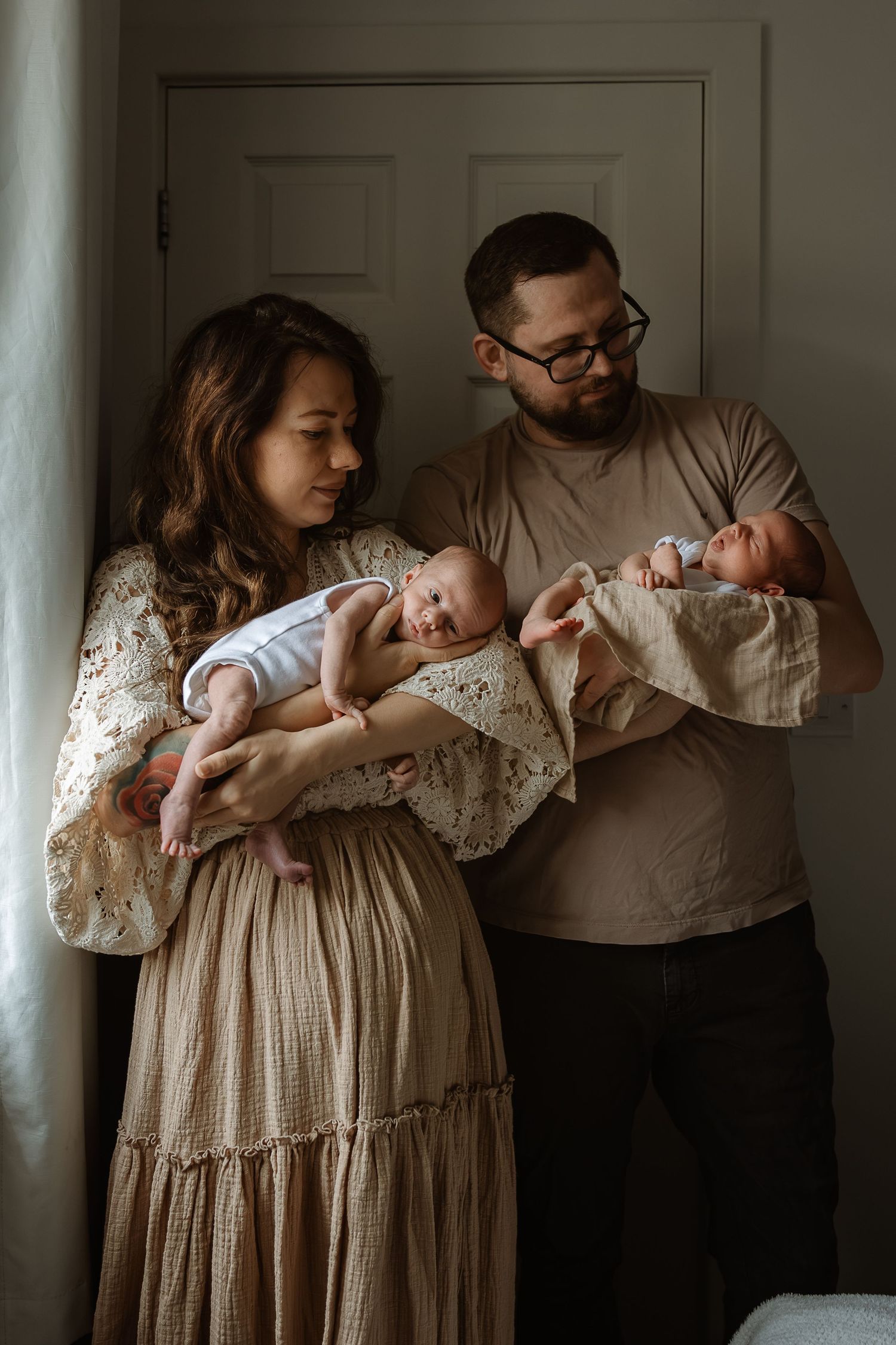 A couple in neutral tones holds their newborn twins in a tender indoor portrait with soft natural lighting.