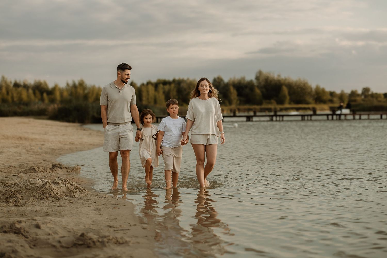 Family wearing white outfits walks along a serene lakeside beach during golden hour.