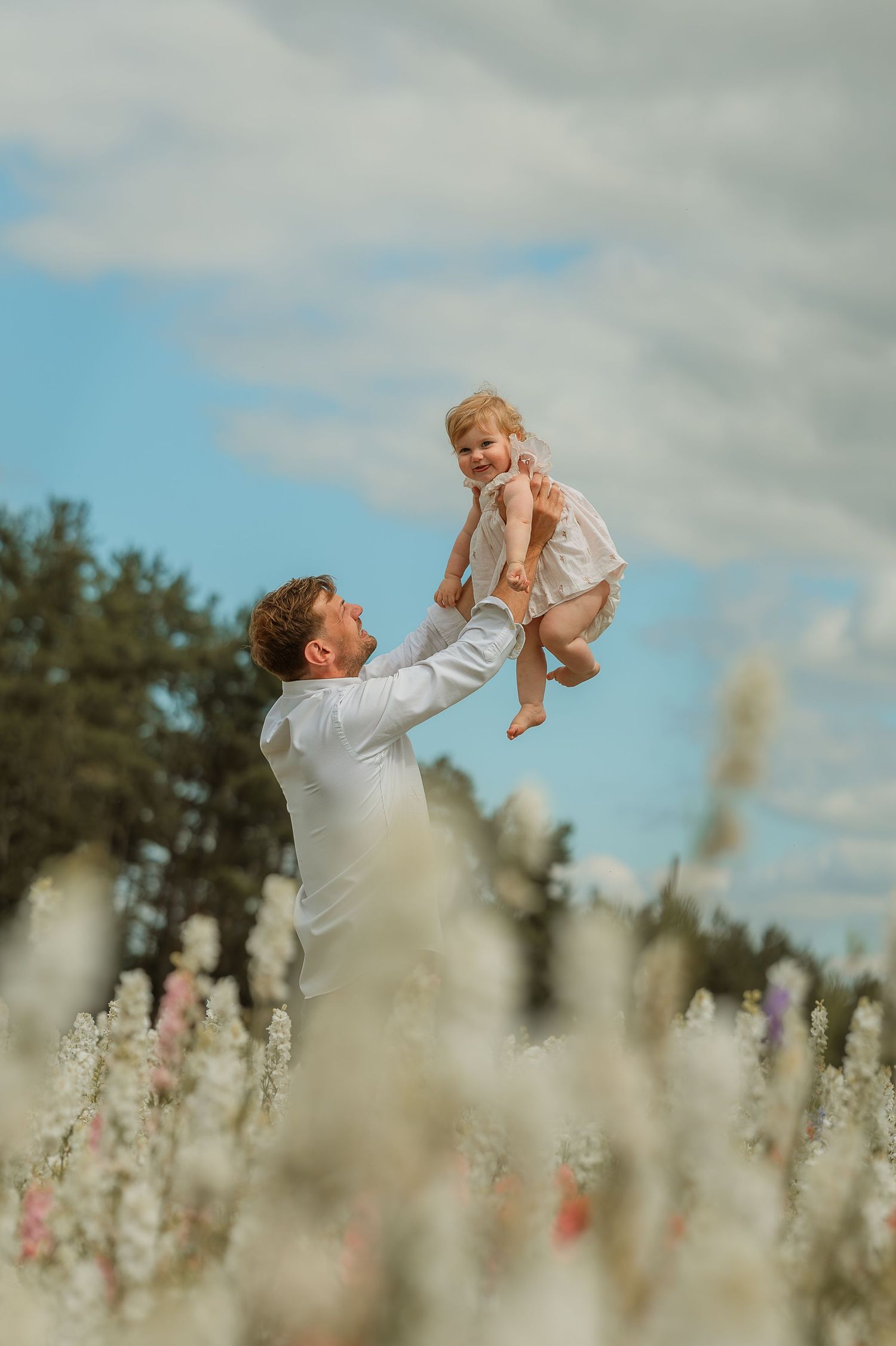 A parent lifts a small child up in the air while standing in a wildflower field on a cloudy summer day.