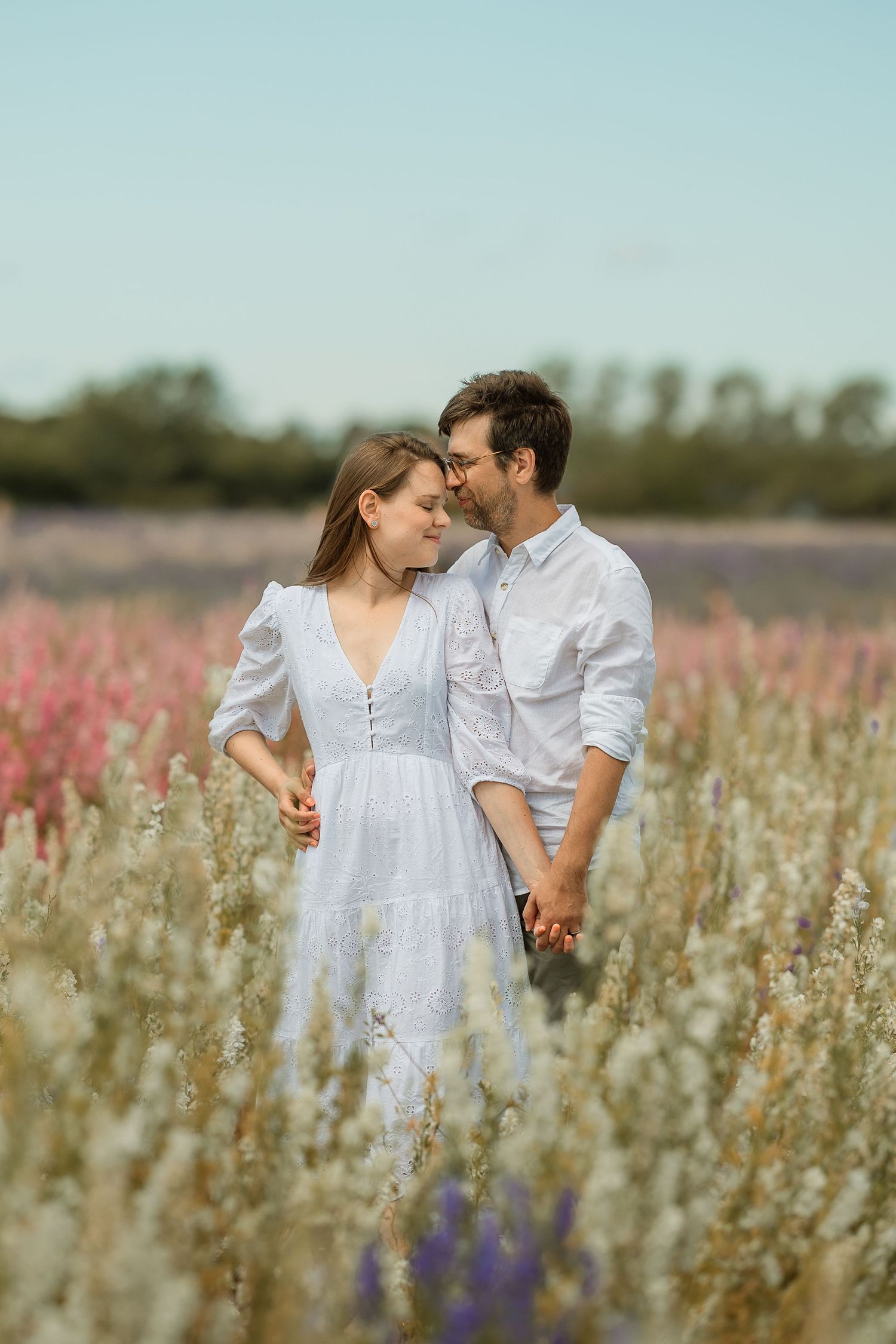 Romantic couple embracing in a blooming wildflower field, woman in white dress, man in white shirt, soft natural light.