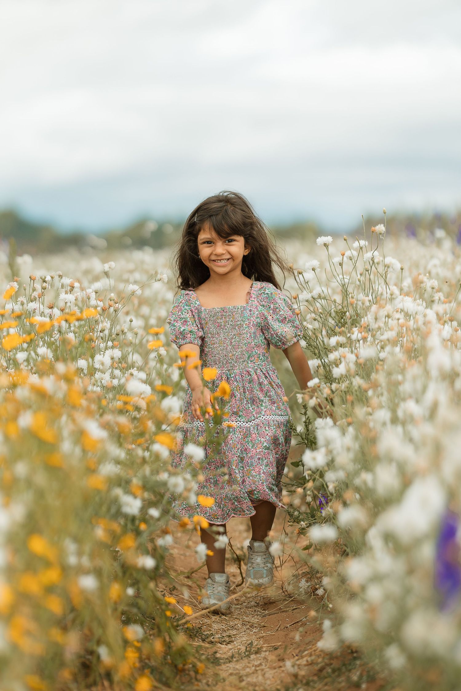 A smiling young girl in a floral dress runs through a vibrant field of white and yellow wildflowers under a cloudy sky.